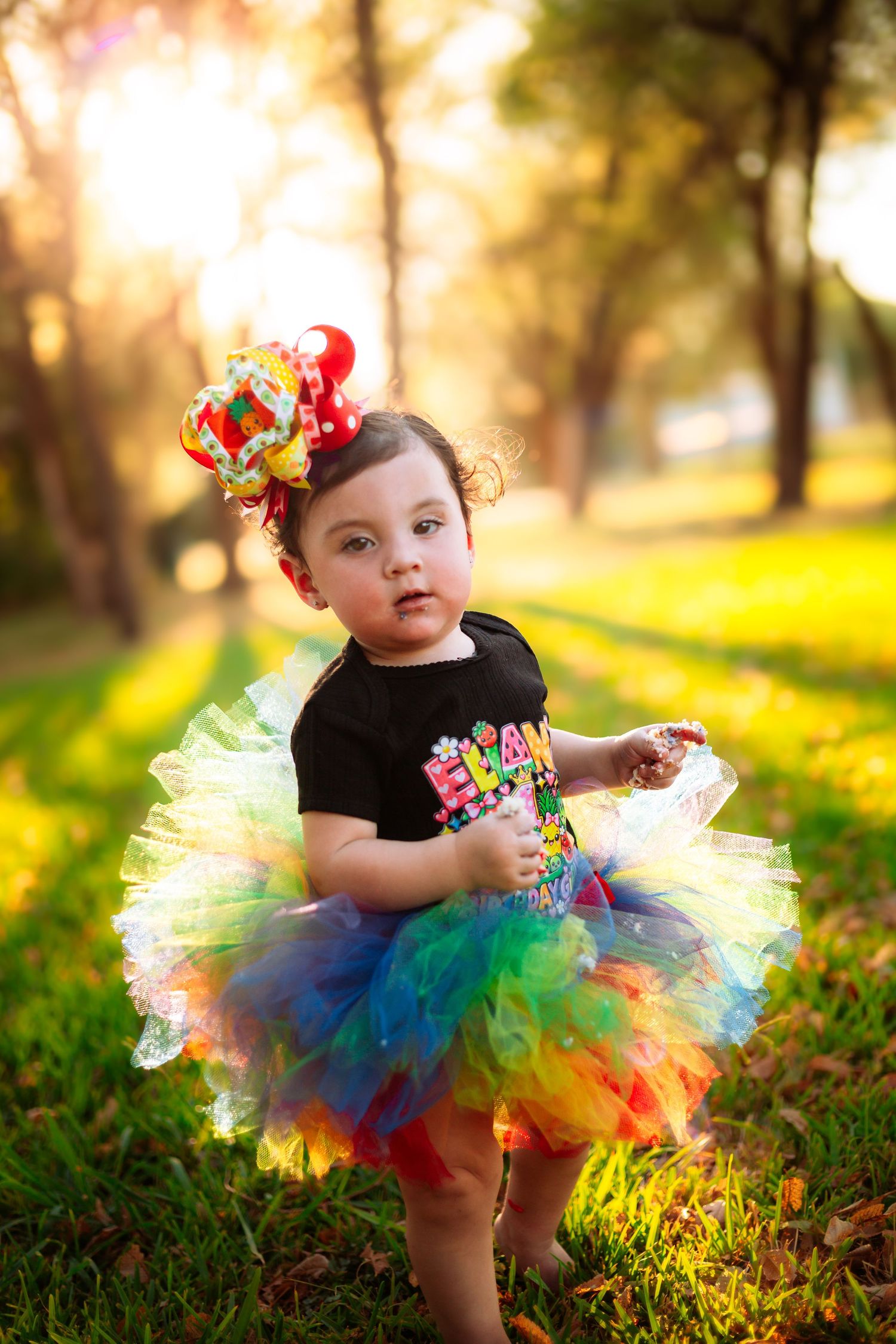 Toddler girl in rainbow tutu skirt and colorful bow headband standing on green grass in sunny park.