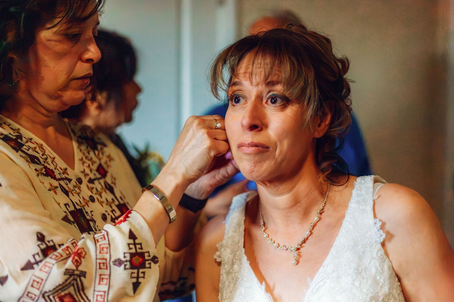 A bride in a white dress gets her earring adjusted by an older woman during wedding preparations.