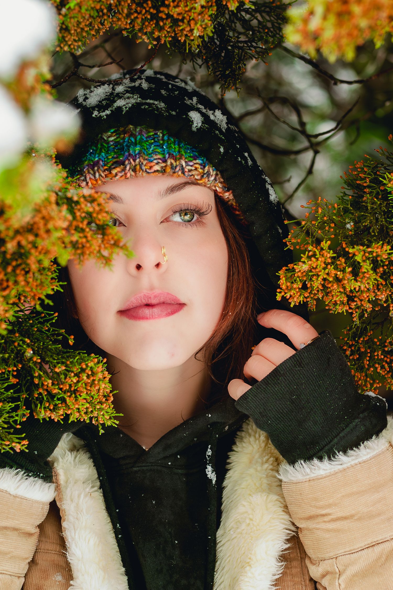 Redhead woman in knit beanie and winter jacket peers through snow-dusted orange and green pine branches.