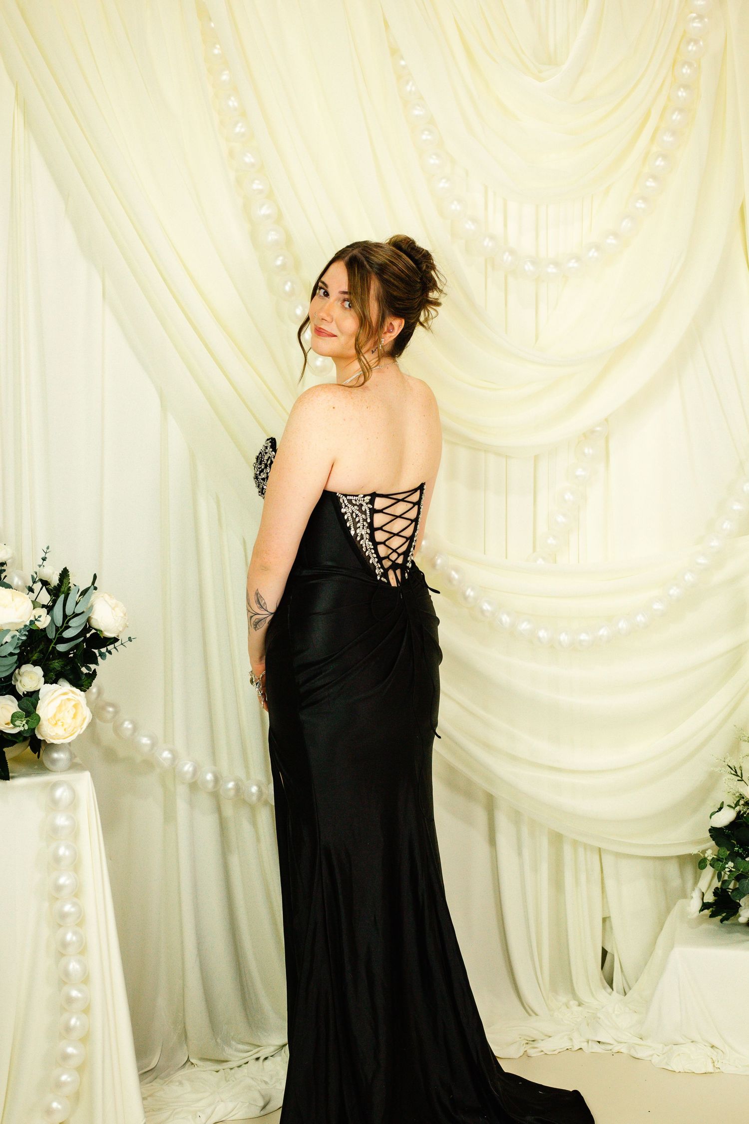 Woman in elegant black strapless lace-up back gown posing in front of white draped fabric backdrop.