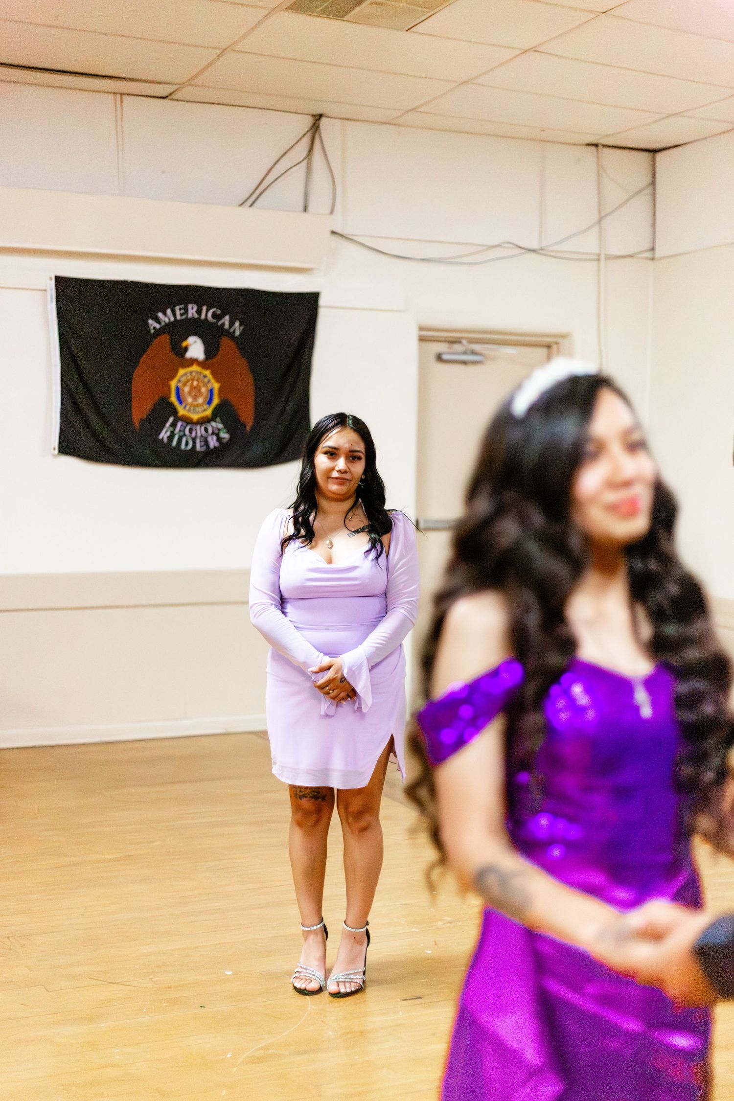Two young women in purple and lavender dresses pose in a decorated hall with an American Riders flag on the wall.