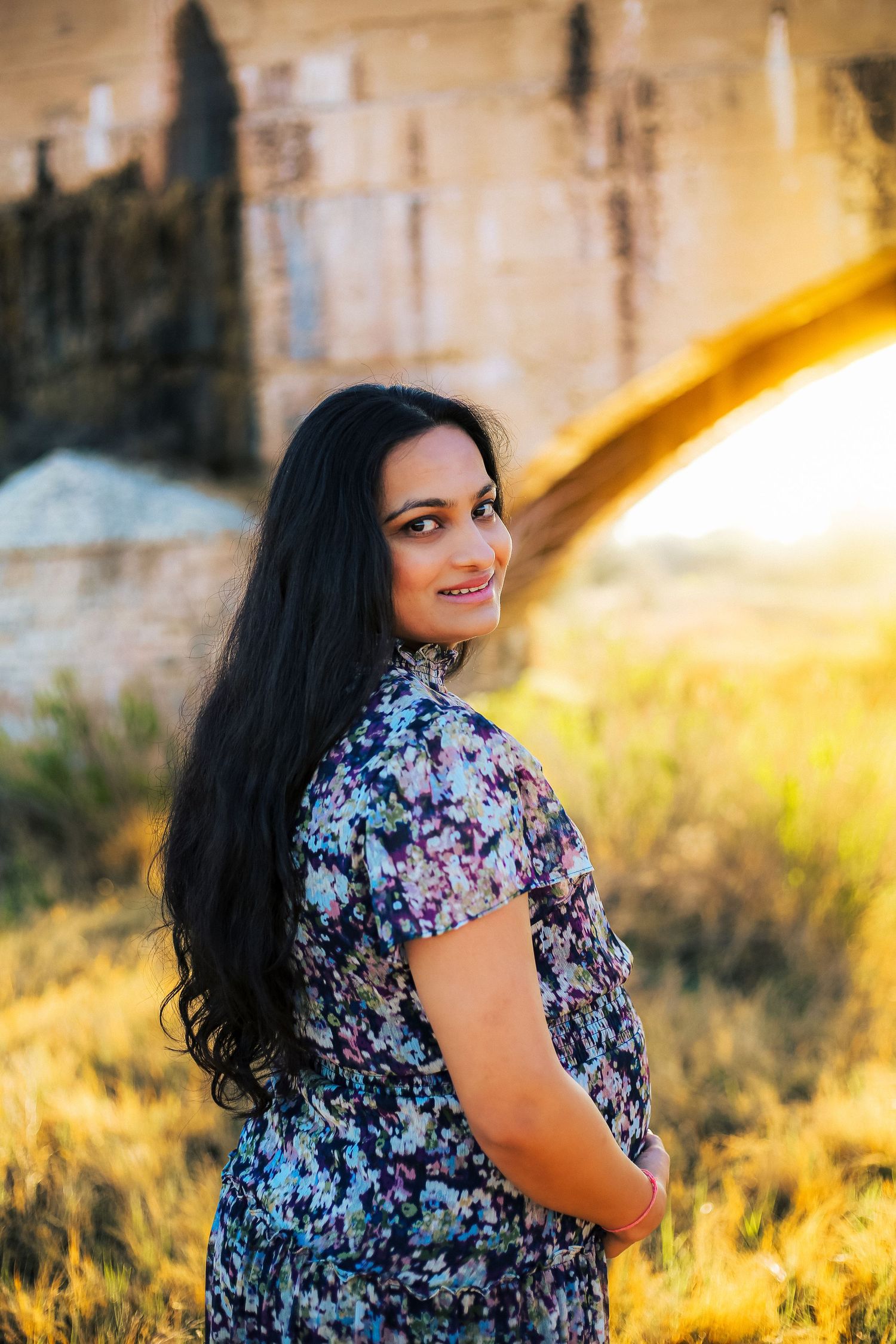 Smiling woman with long dark hair in floral dress poses outdoors at golden hour near rustic stone structure.