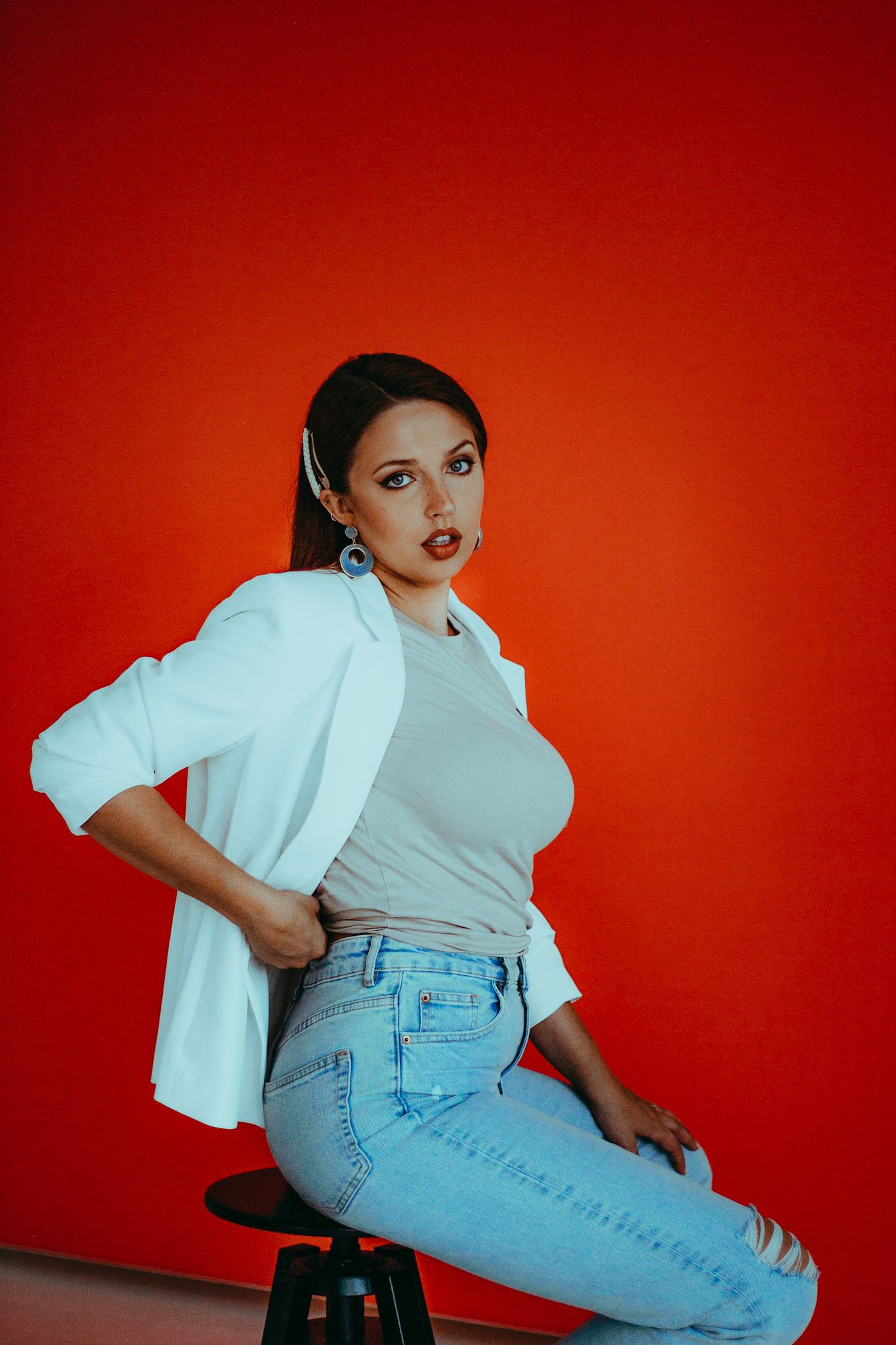 Stylish woman in white blazer and ripped jeans sits on stool against vibrant red wall.