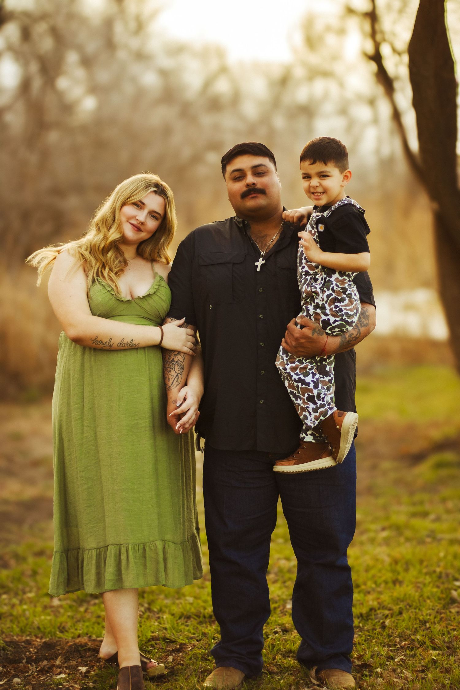 Happy family portrait with tattooed parents and toddler son in golden autumn park setting.