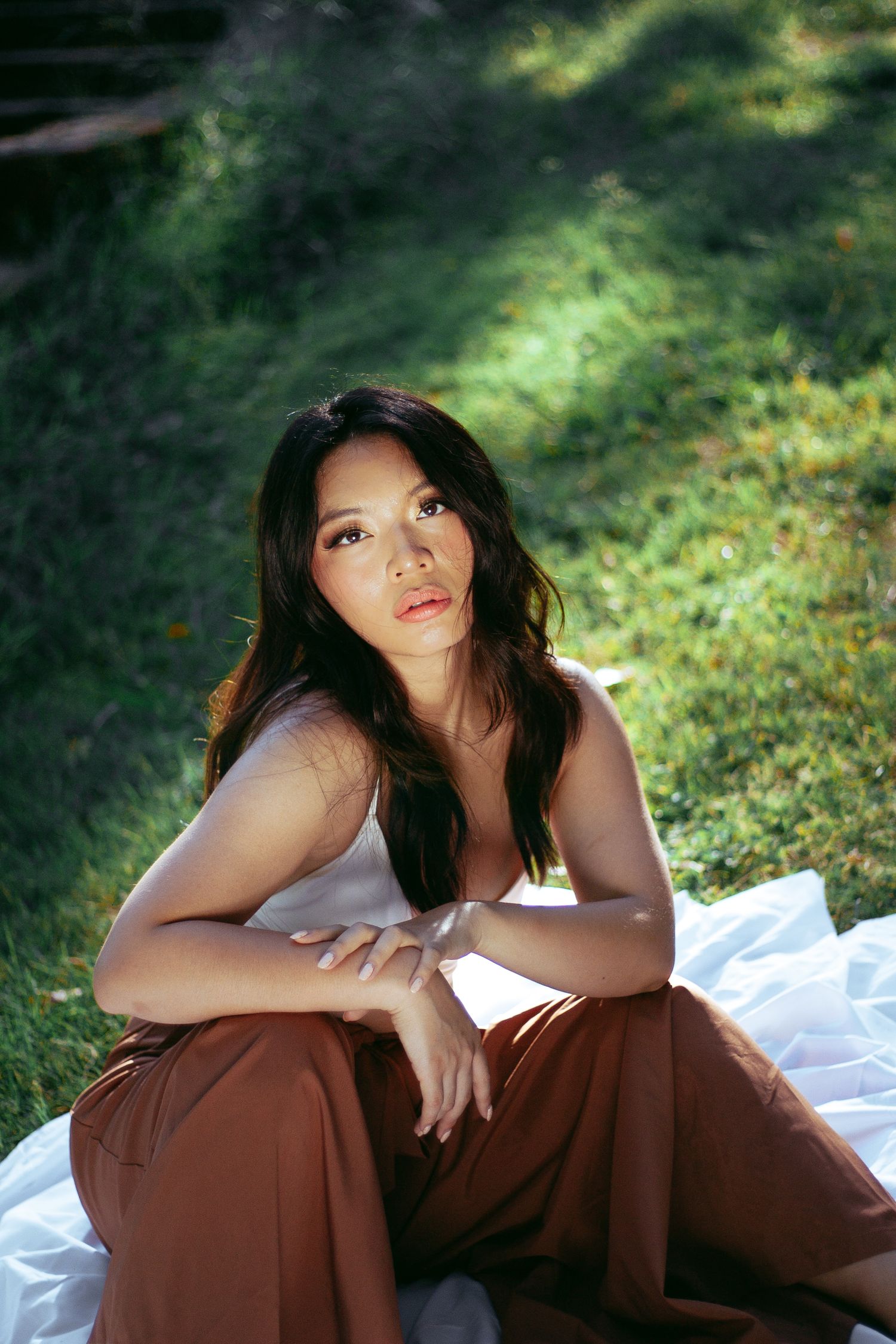 Woman in white top and brown pants sitting on grass in dappled sunlight.