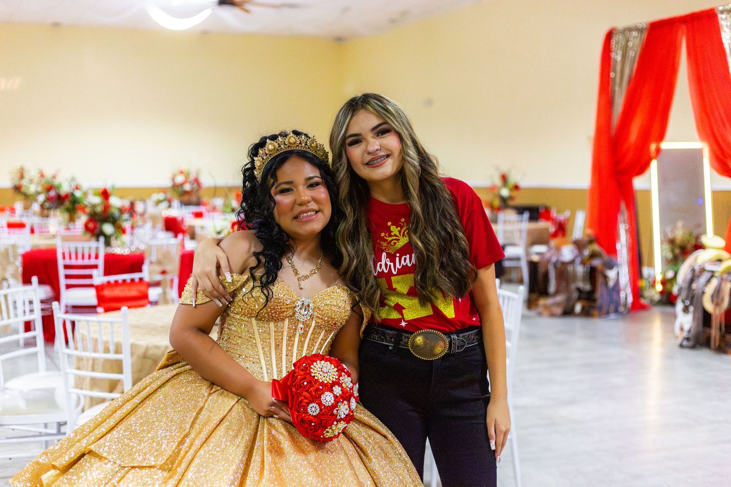 Two young women pose together at a quinceañera celebration, one wearing a gold ball gown and holding a red bouquet.