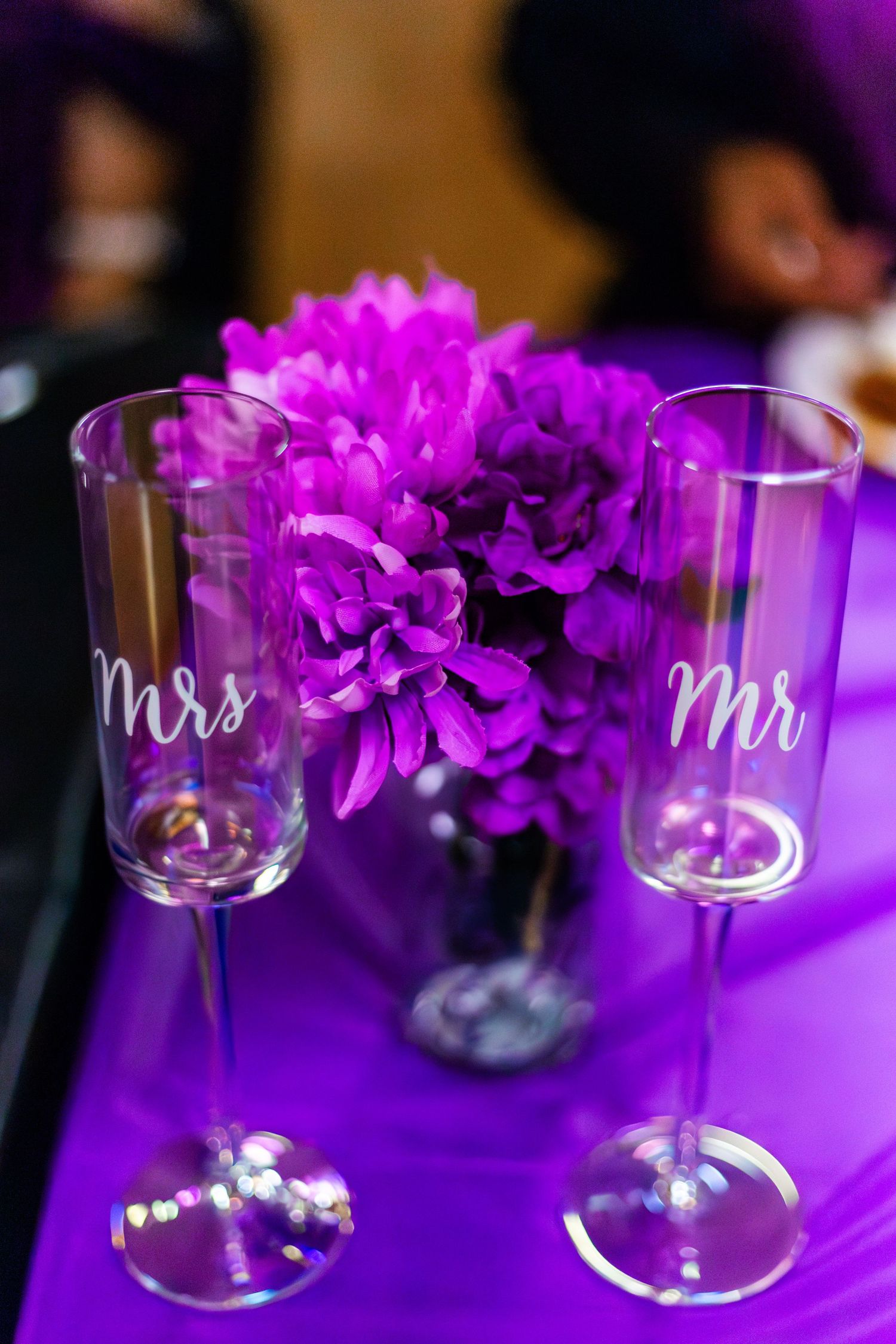 Mr. and Mrs. champagne flutes on purple tablecloth with vibrant magenta floral centerpiece at wedding reception.