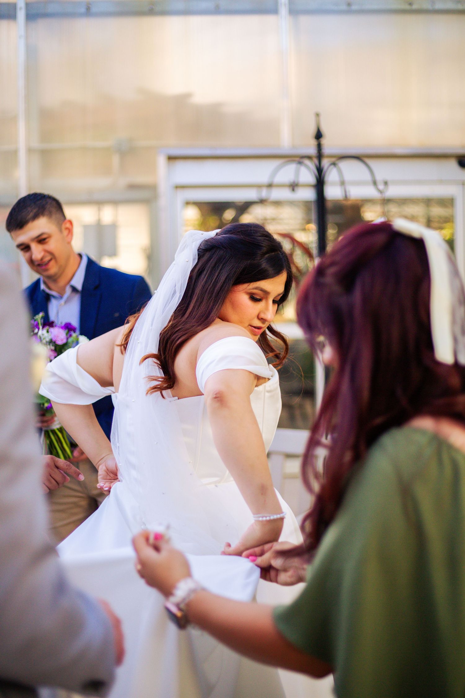 Bride in off-shoulder white gown gets dress adjusted by attendants while groom waits in greenhouse venue.
