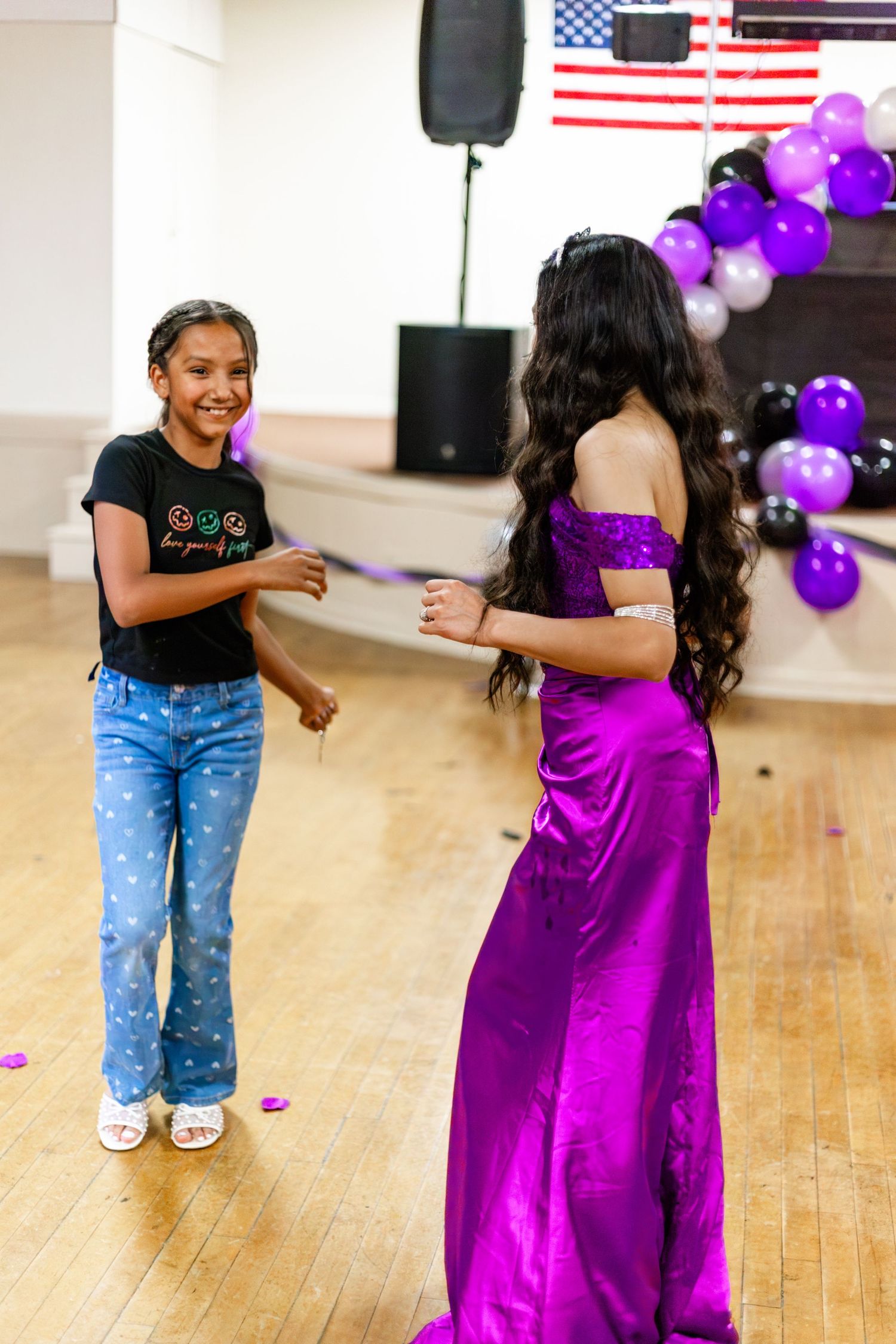 Two girls dancing at a quinceañera party with purple balloons and an American flag in the background.