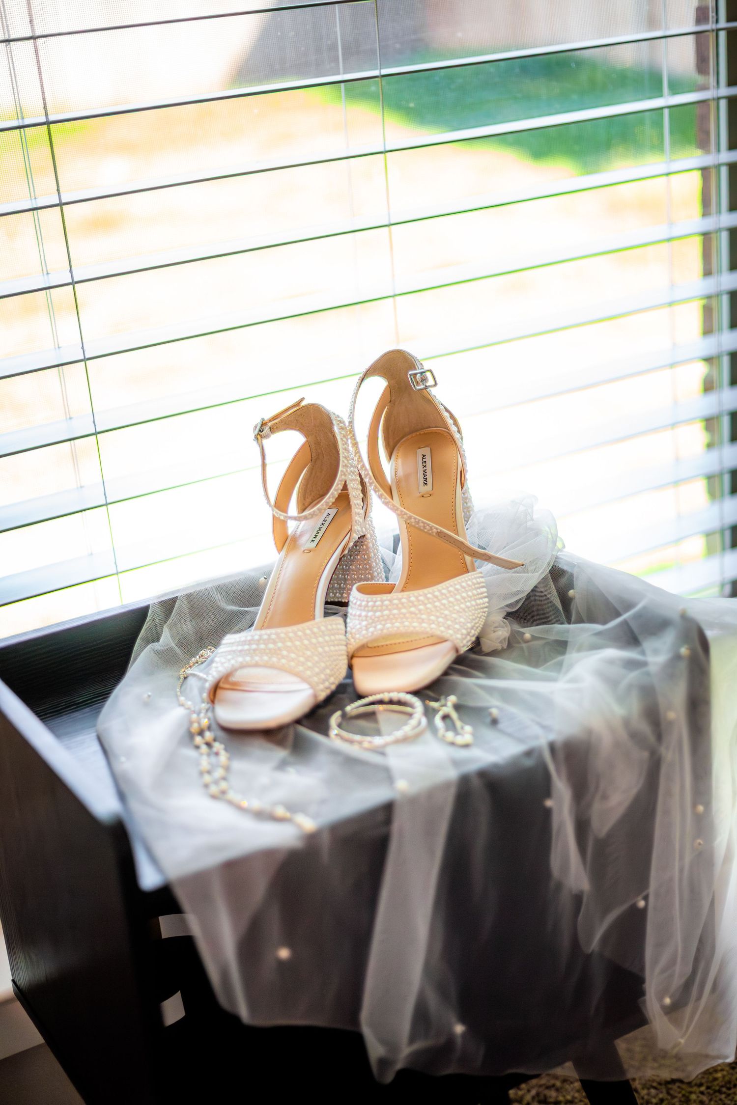 Elegant white bridal heels with ankle straps displayed on a dark surface with pearl jewelry and veil near a sunlit window.