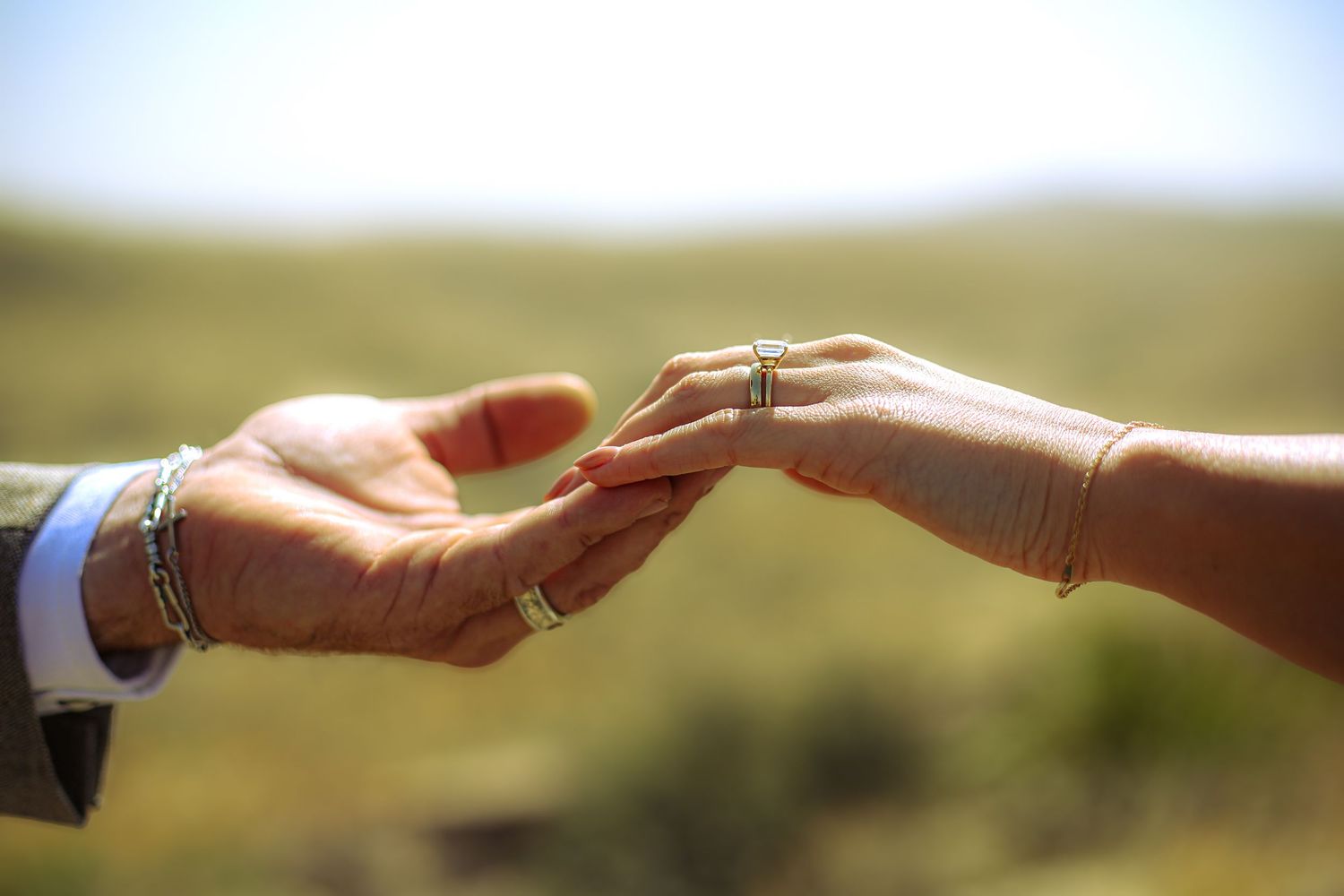 Two hands touching outdoors, adorned with rings and bracelets, symbolizing a romantic engagement or wedding moment.