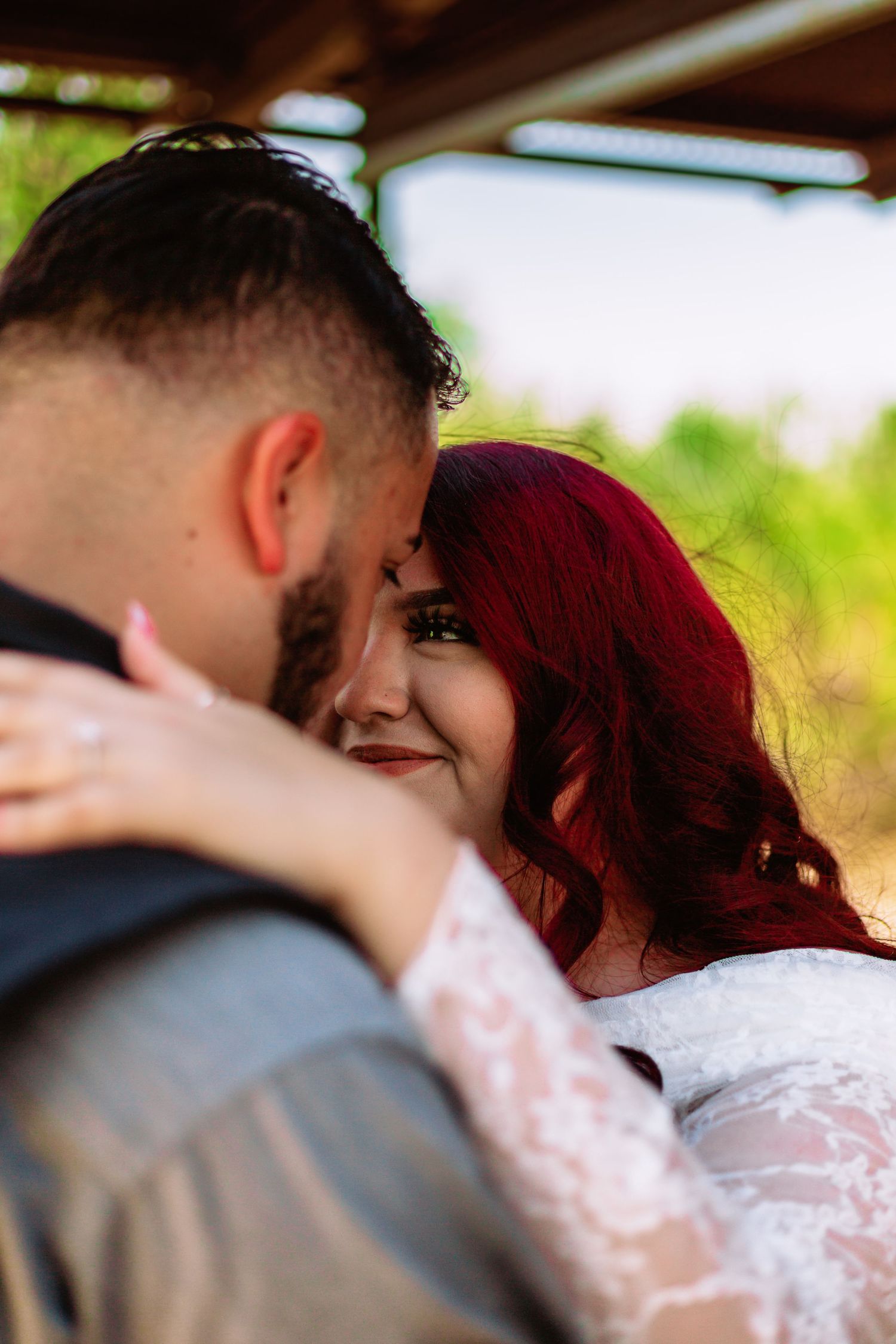 A bride with red hair in lace dress embraces her bearded groom outdoors in a romantic wedding portrait.
