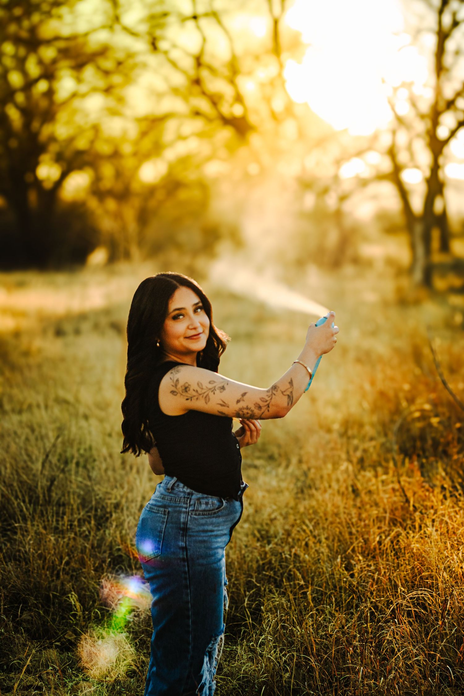 A young woman with floral arm tattoos poses in a sunlit golden field during a warm outdoor portrait.