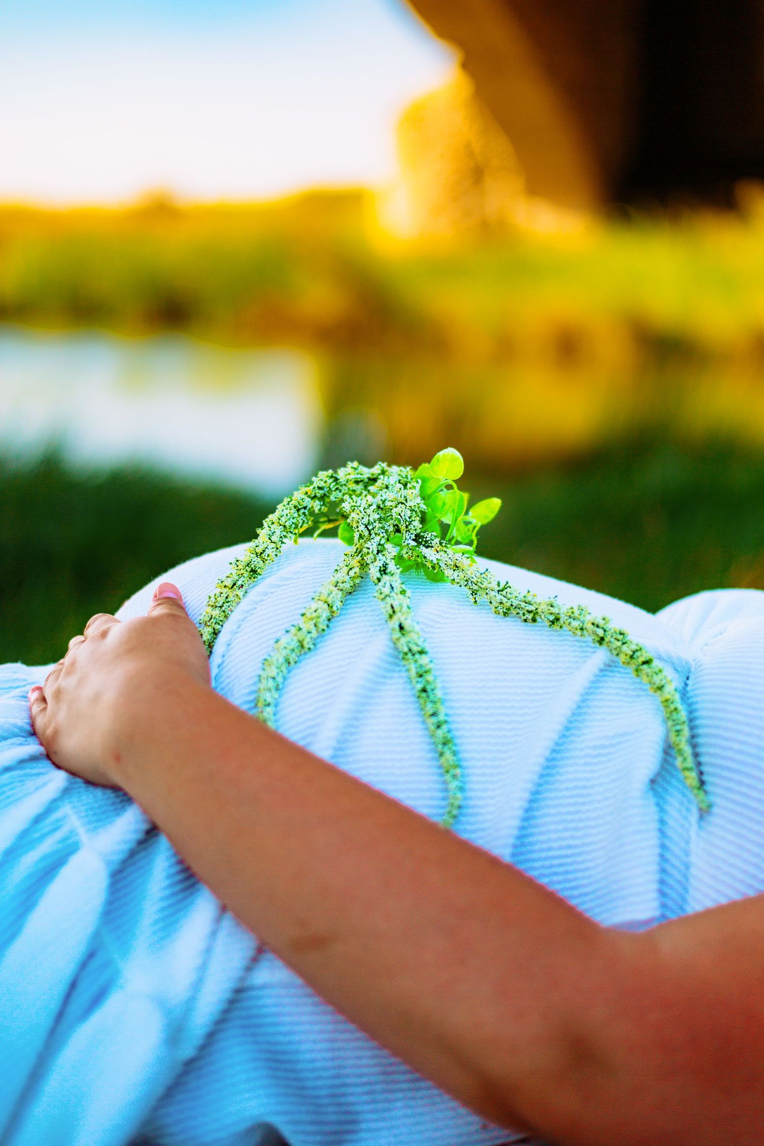Pregnant woman in blue dress resting outdoors holding small white wildflowers on her baby bump near a serene river.