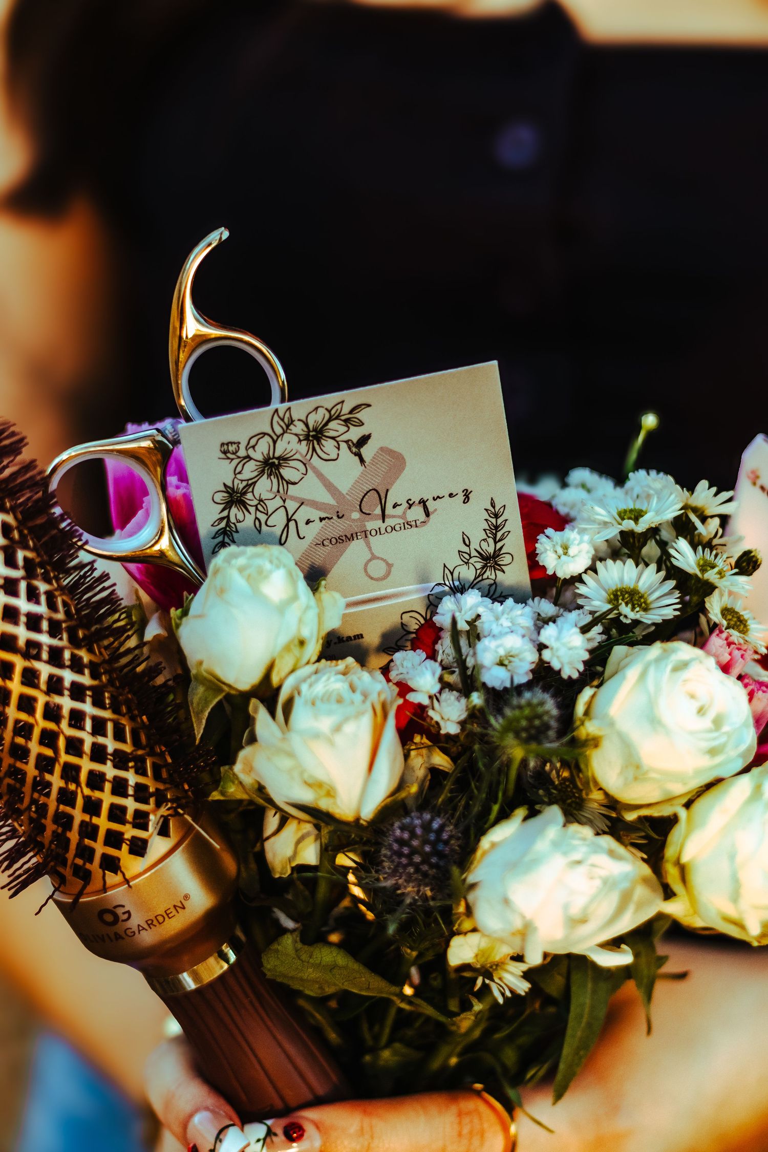 A florist holds a bouquet of white roses and wildflowers with gold scissors and a business card tucked inside.