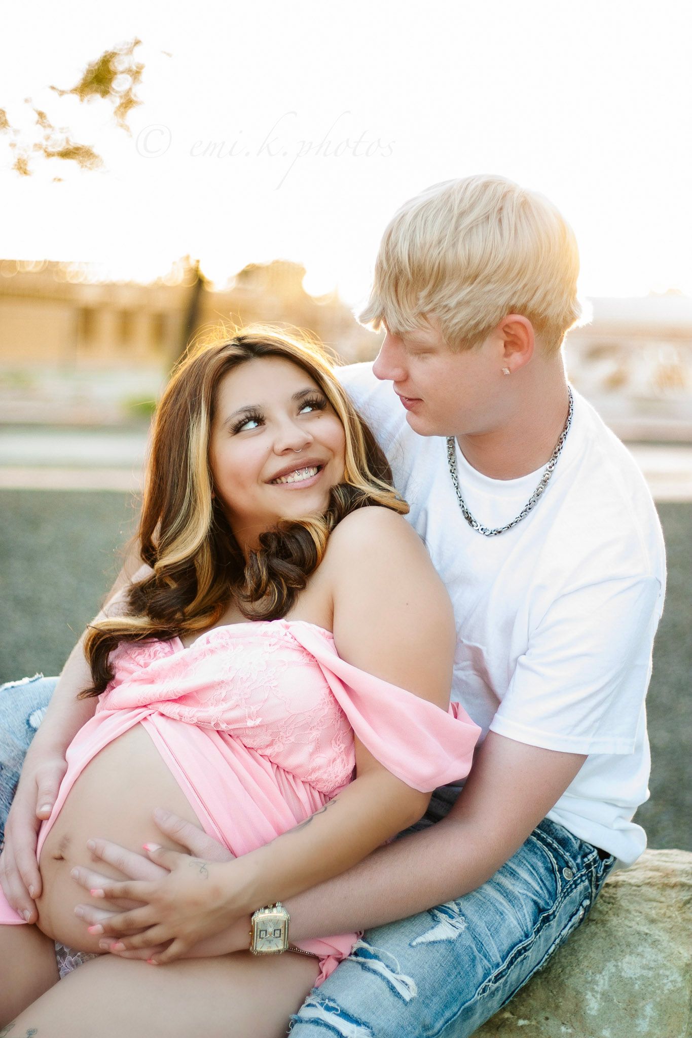 Smiling pregnant woman in pink top sitting with blonde partner at golden hour outdoors.