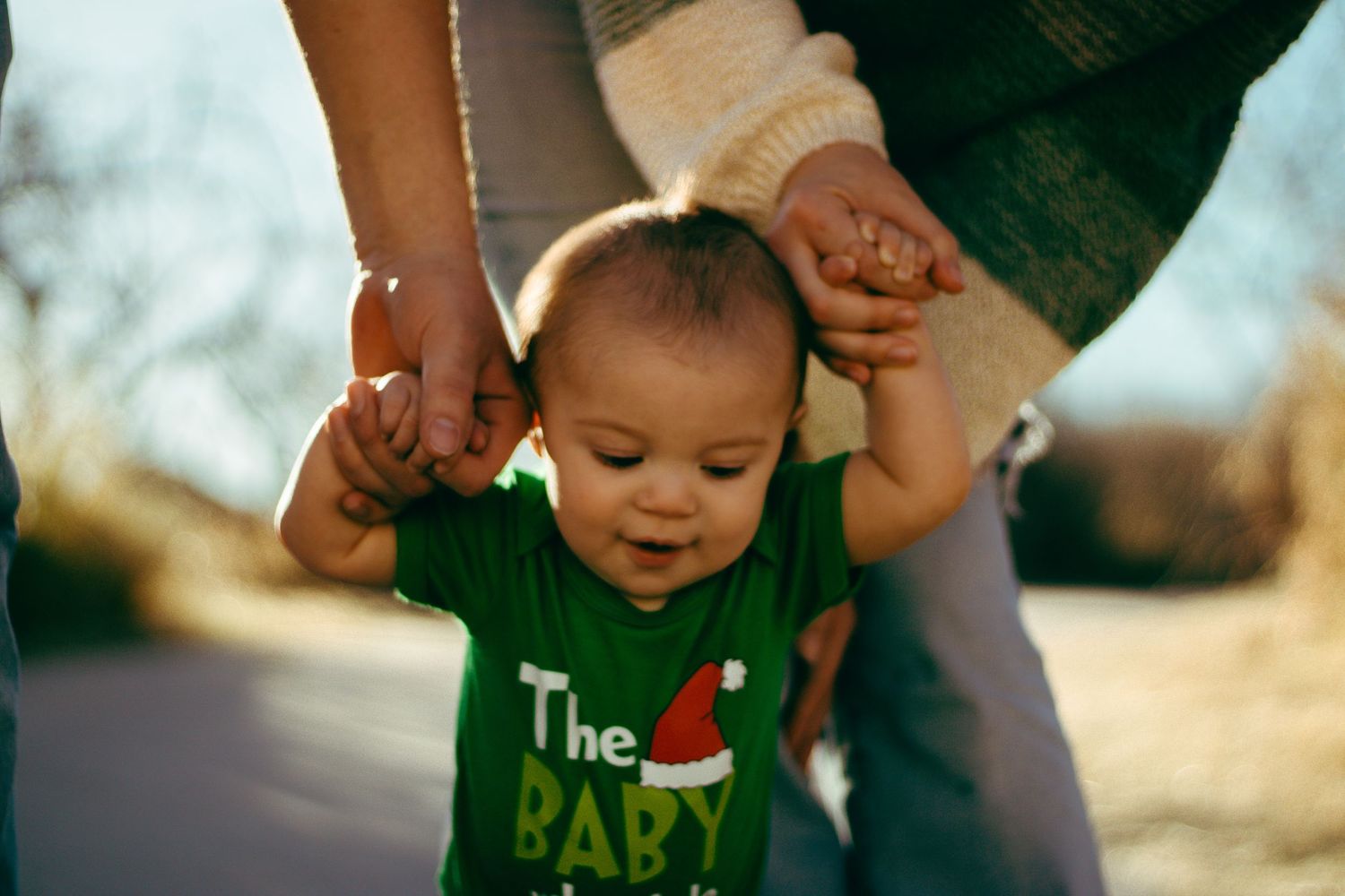 Baby in Christmas Grinch onesie taking first steps held by parents.