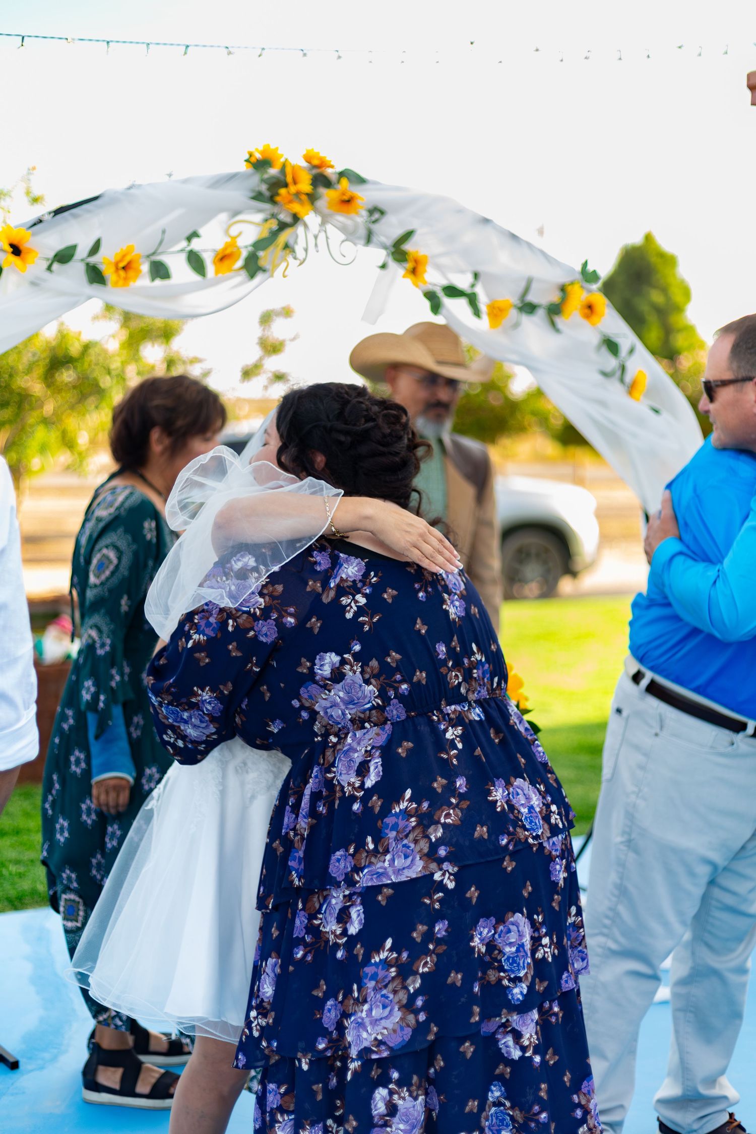 Bride receives warm hug from guest in floral dress under sunflower-decorated wedding arch.