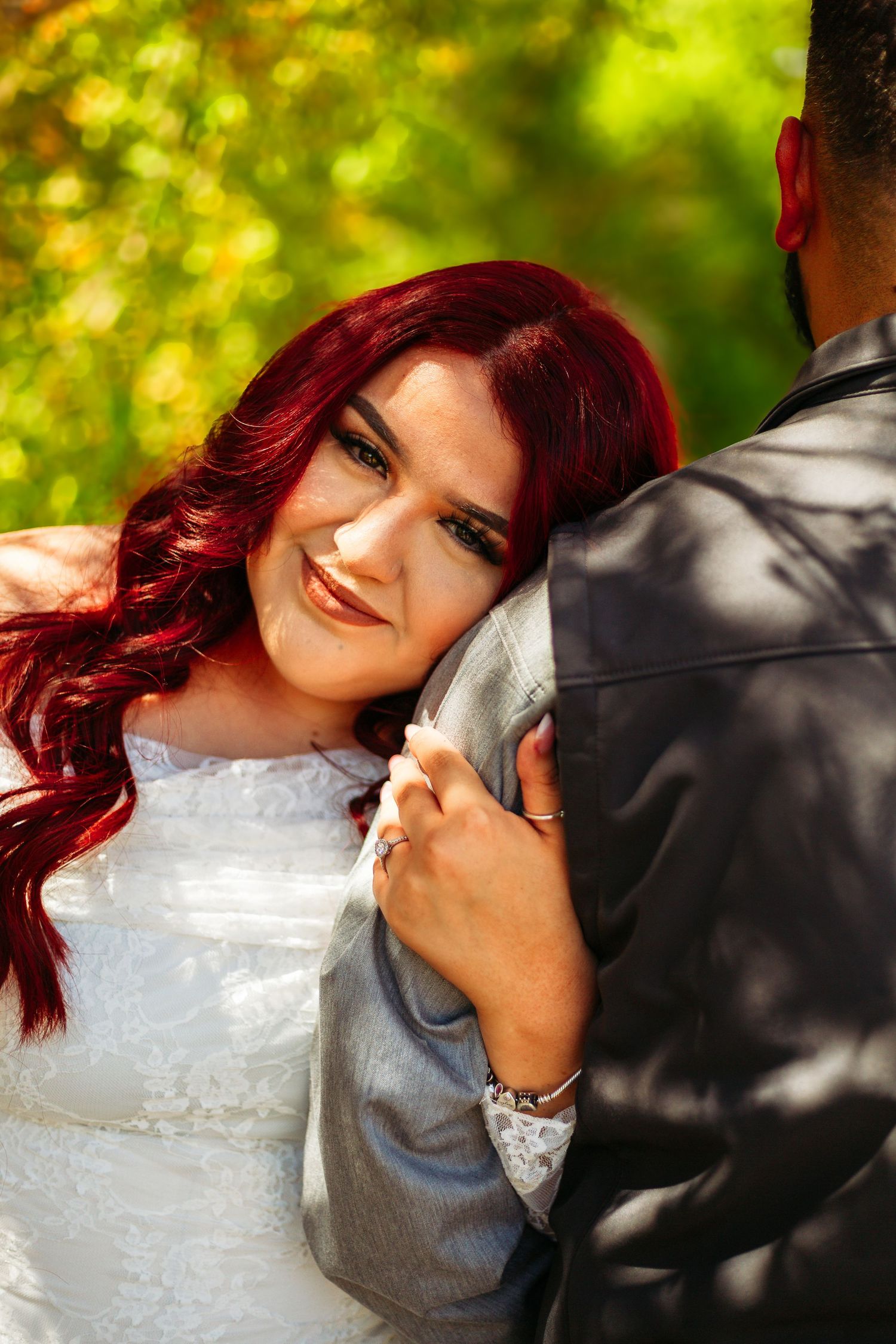 Smiling bride with red hair in white lace dress embracing groom in gray suit outdoors with green foliage background.