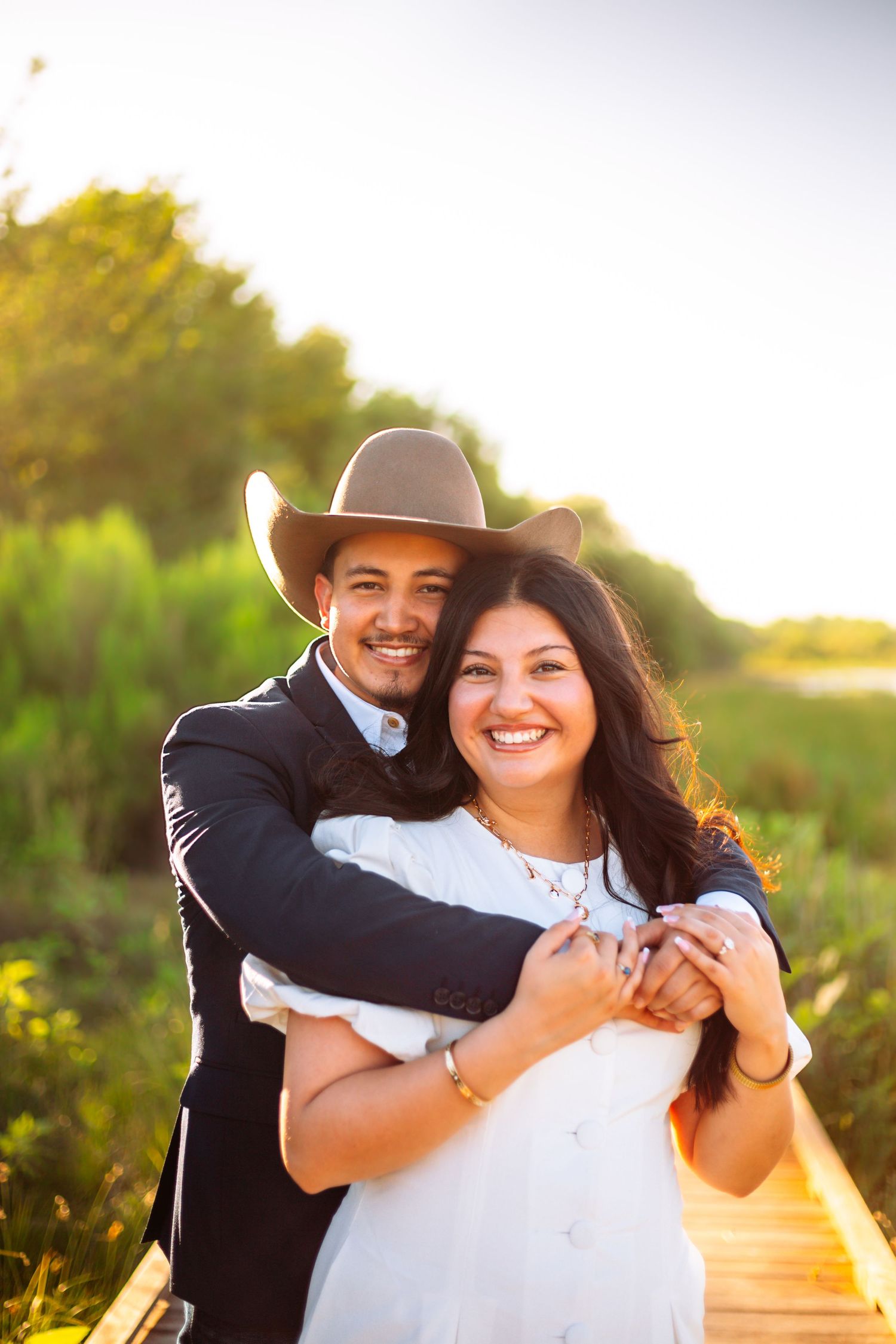 Happy couple embracing outdoors at golden hour, man in cowboy hat hugging woman in white dress on wooden boardwalk.