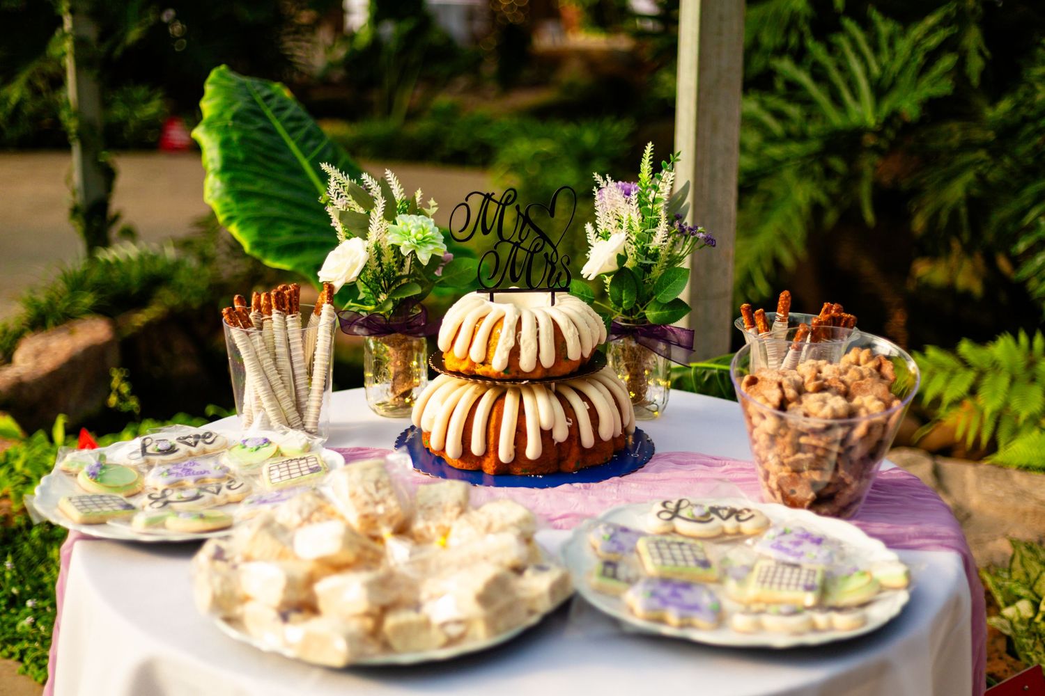 Elegant outdoor dessert table with bundt cake, assorted sweets, and floral decor at a garden celebration.