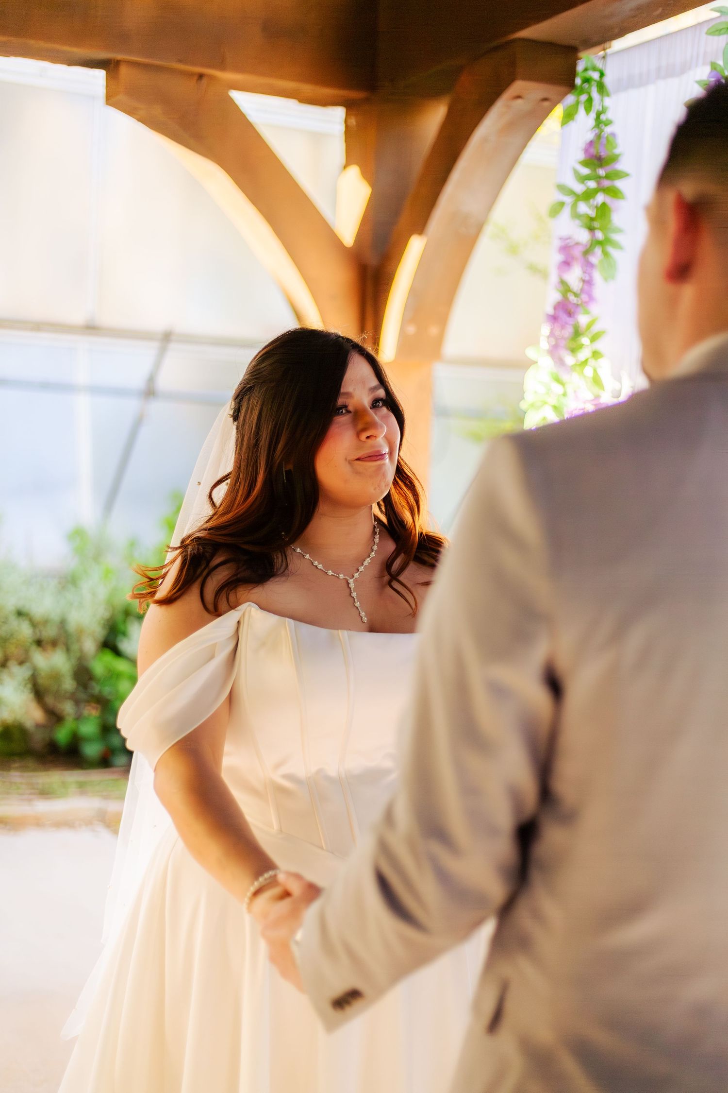 Bride in white off-shoulder dress smiles at groom during intimate wedding ceremony under wooden arch with floral decor.