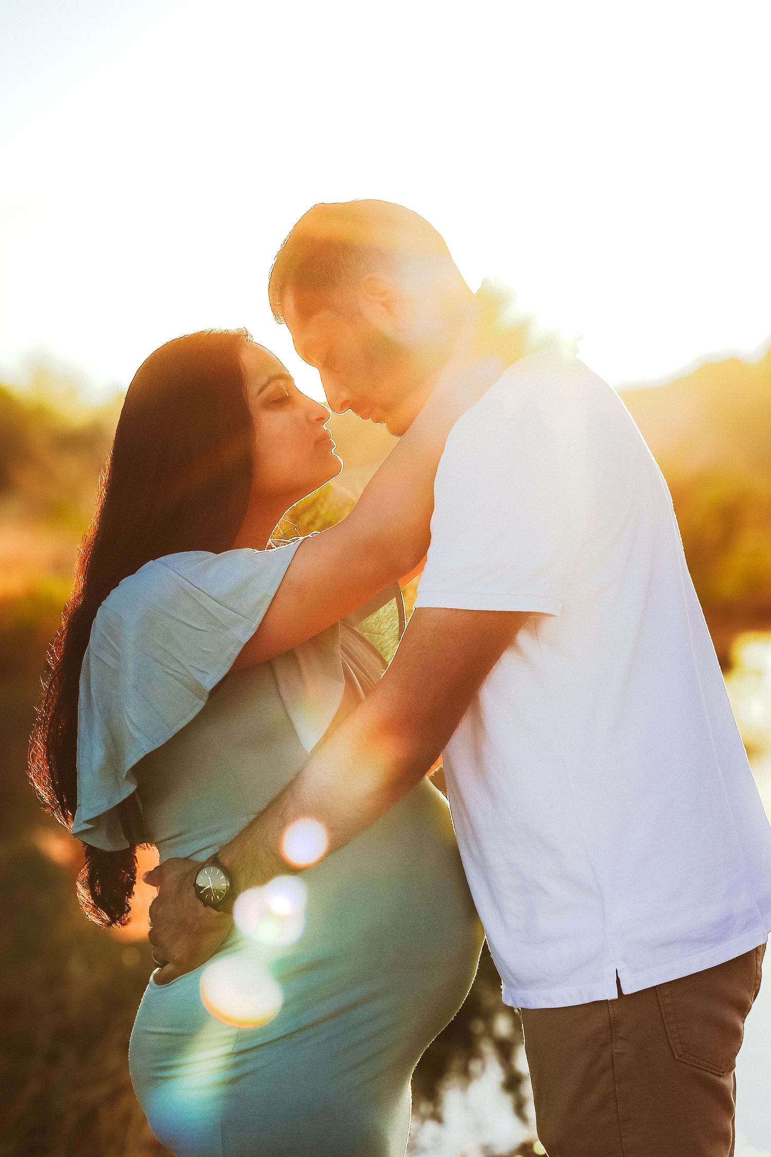 Pregnant couple sharing a kiss in warm golden sunset light outdoors.