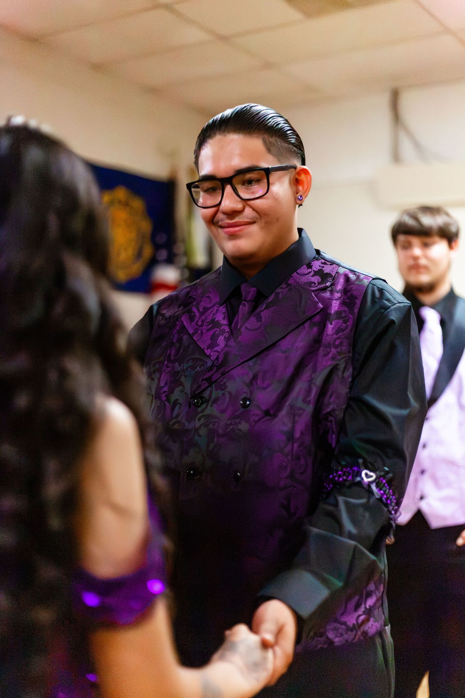 Young man in purple floral tuxedo jacket and glasses smiling at prom event.