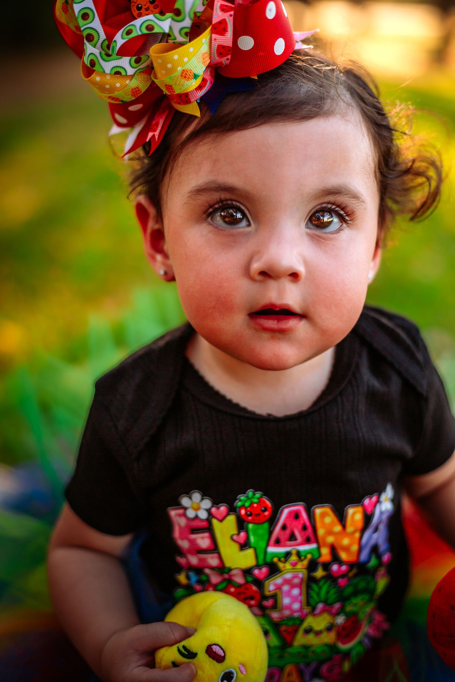 Adorable baby girl with big brown eyes wearing a colorful bow headband and Eliana shirt holding a toy.