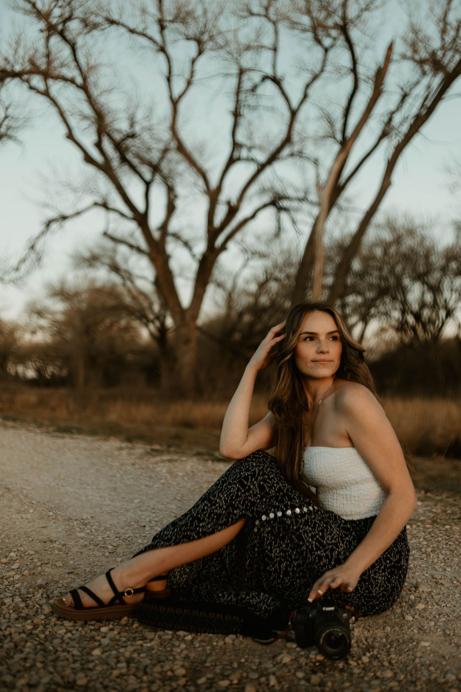 Young woman in white top and dark floral skirt sitting on dirt road holding camera at dusk.