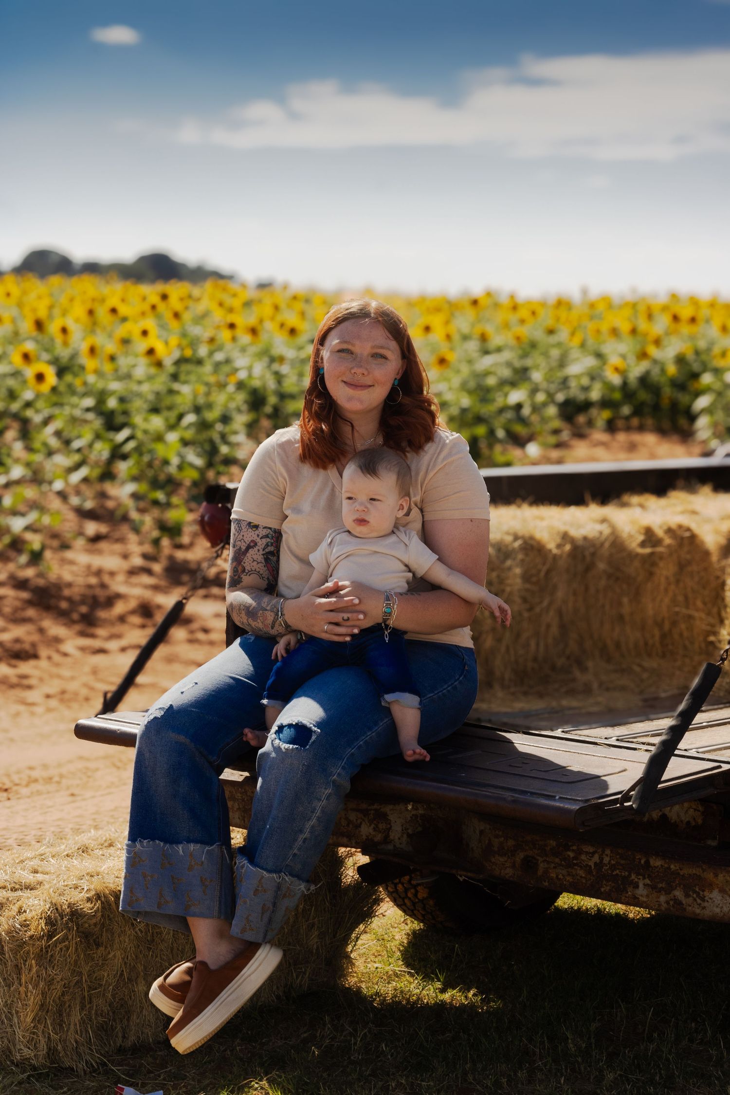 Red-haired mother holding baby on hay wagon in front of sunflower field.