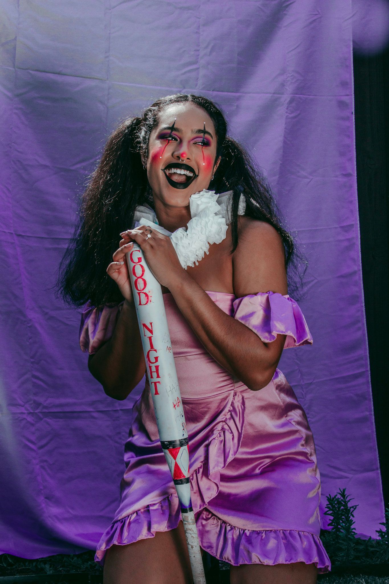 Woman in pink satin dress with clown makeup holding a bat labeled Good Night on purple backdrop.