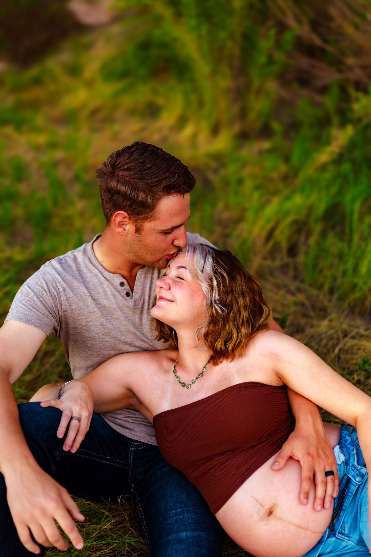 A couple sits in a grassy field, man kissing pregnant woman's head while she smiles and cradles her baby bump.