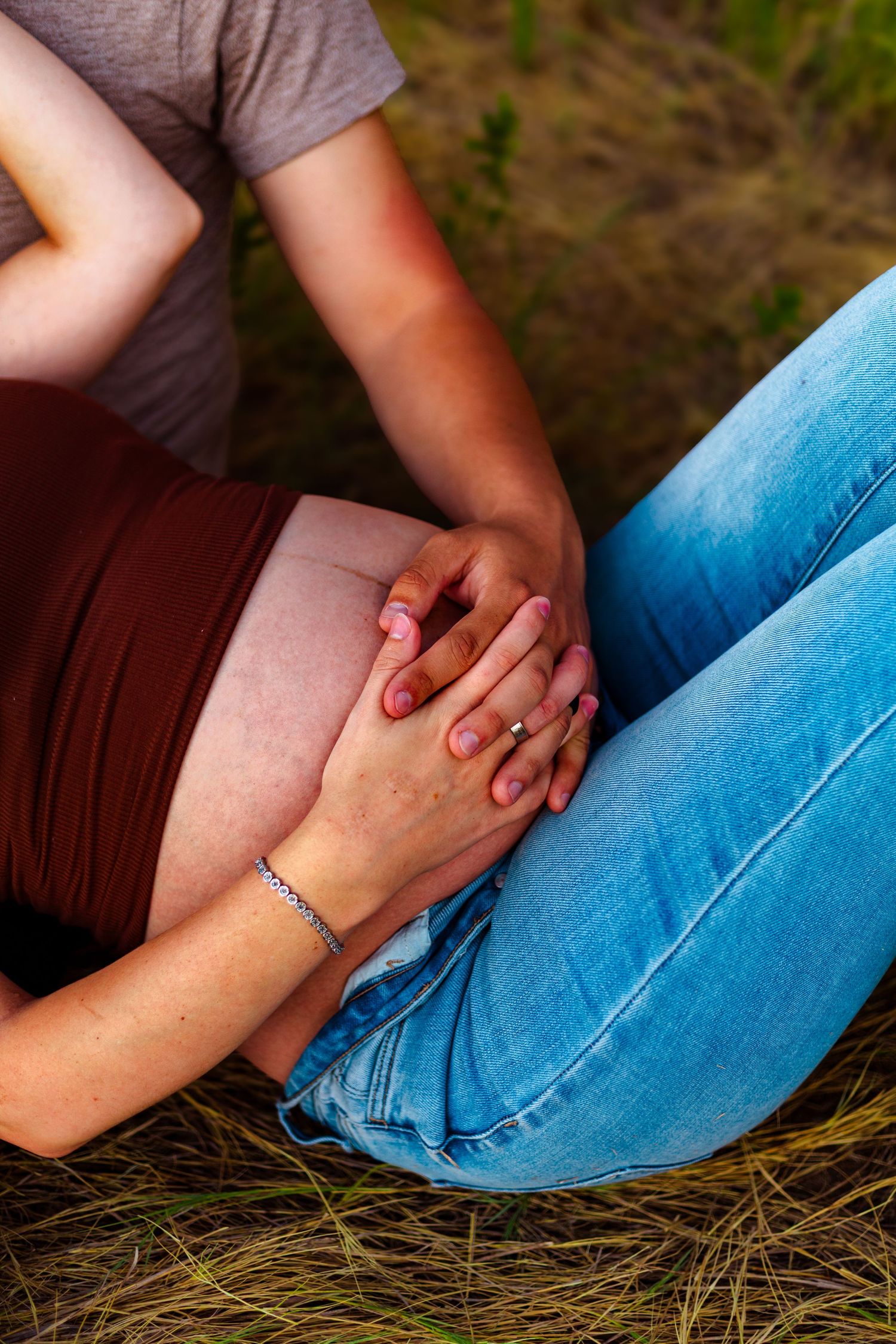 Couple holding hands on pregnant belly outdoors, sitting on grass, woman wearing bracelet and red top.