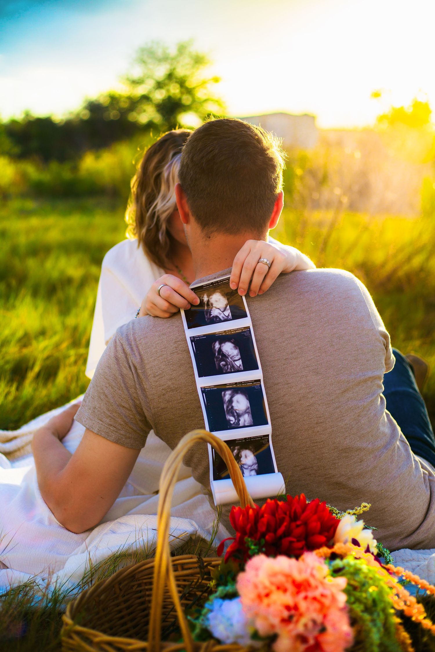 Couple sitting on blanket at sunset holding ultrasound photos, with flower basket nearby, announcing pregnancy outdoors.