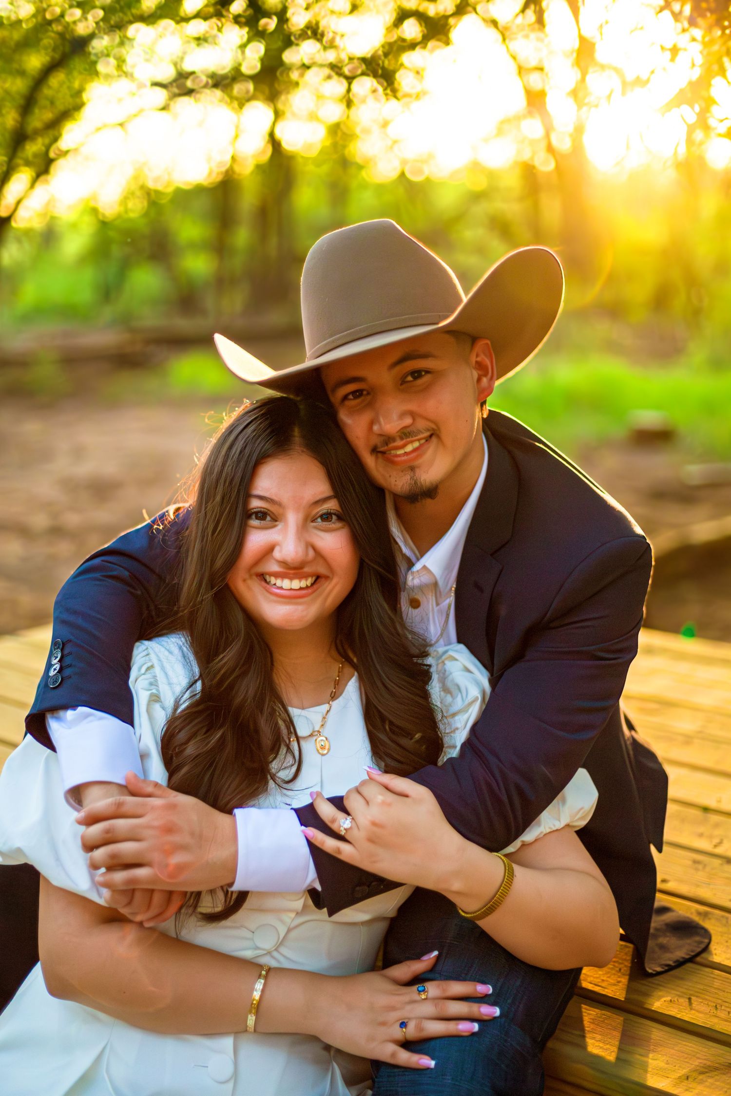 A smiling couple poses outdoors at golden hour, man wearing cowboy hat and navy blazer embracing woman in white dress.