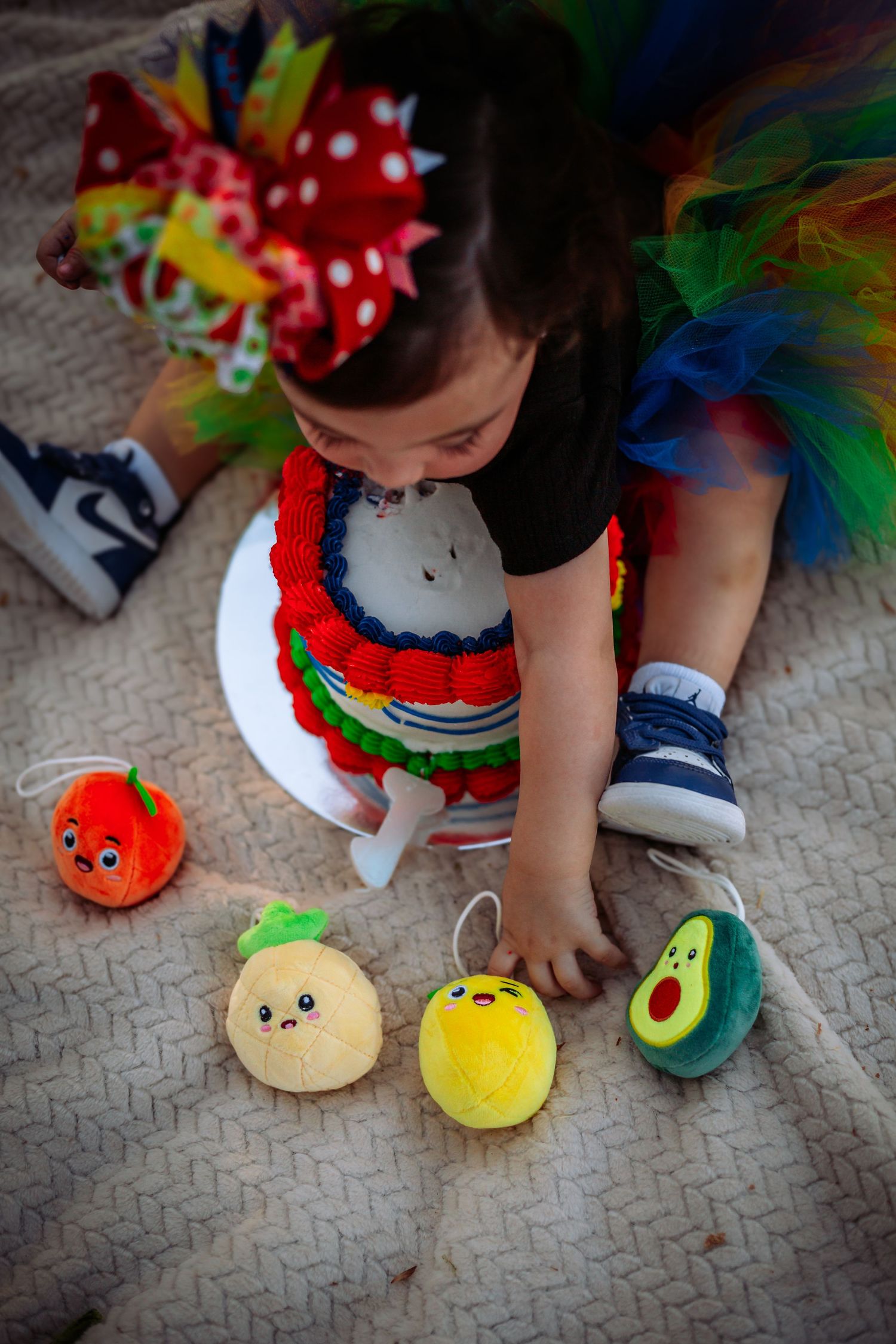 Toddler girl in colorful tutu and bow plays with fruit plush toys on blanket.