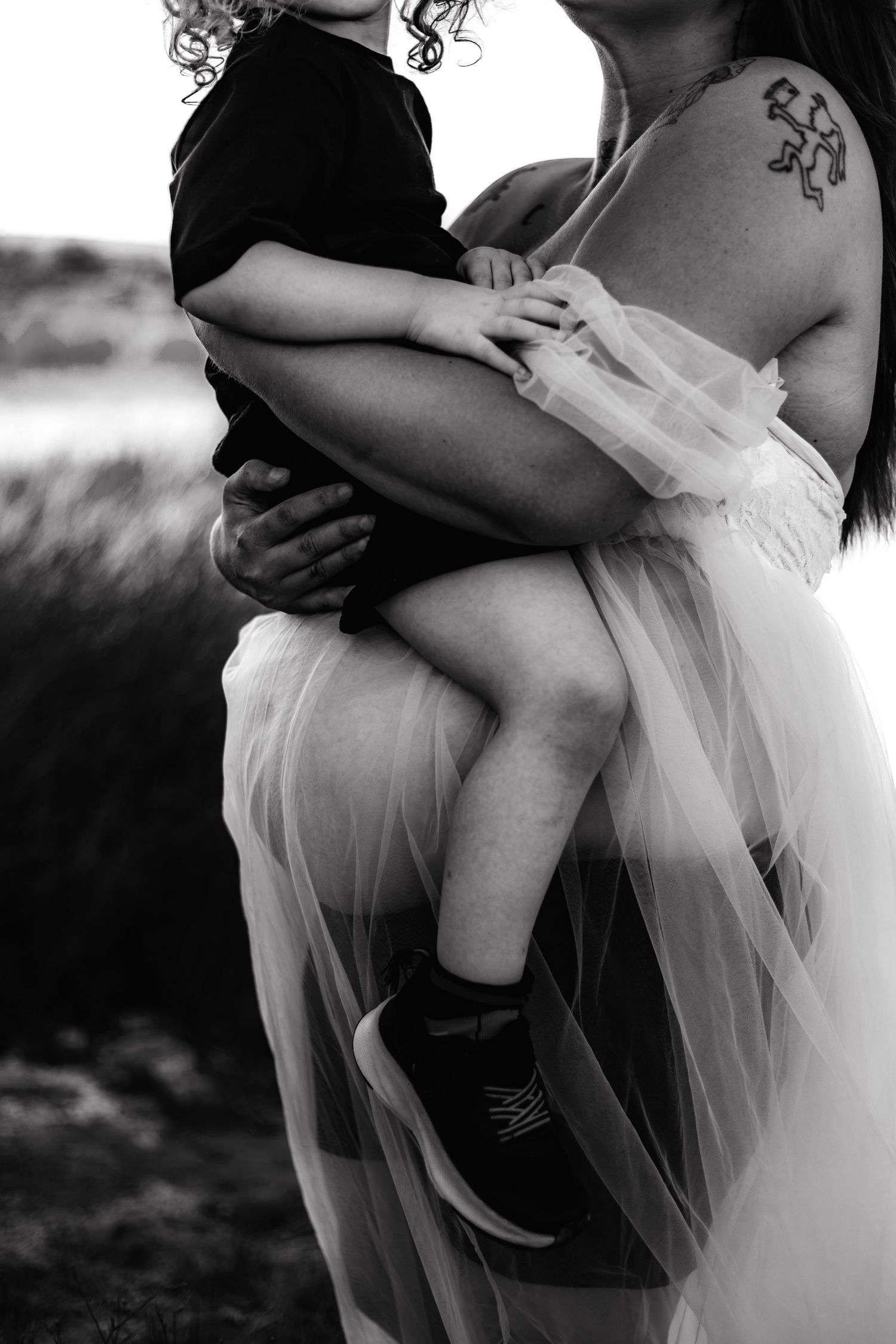 Black and white photo of a mother in a tulle skirt with a tattoo holding a young child outdoors.