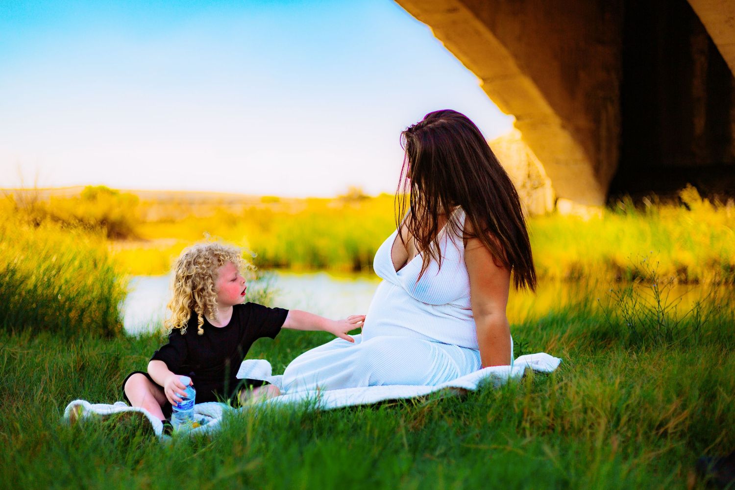 Pregnant woman in white dress sitting on grass with curly-haired child near a bridge on a sunny day.