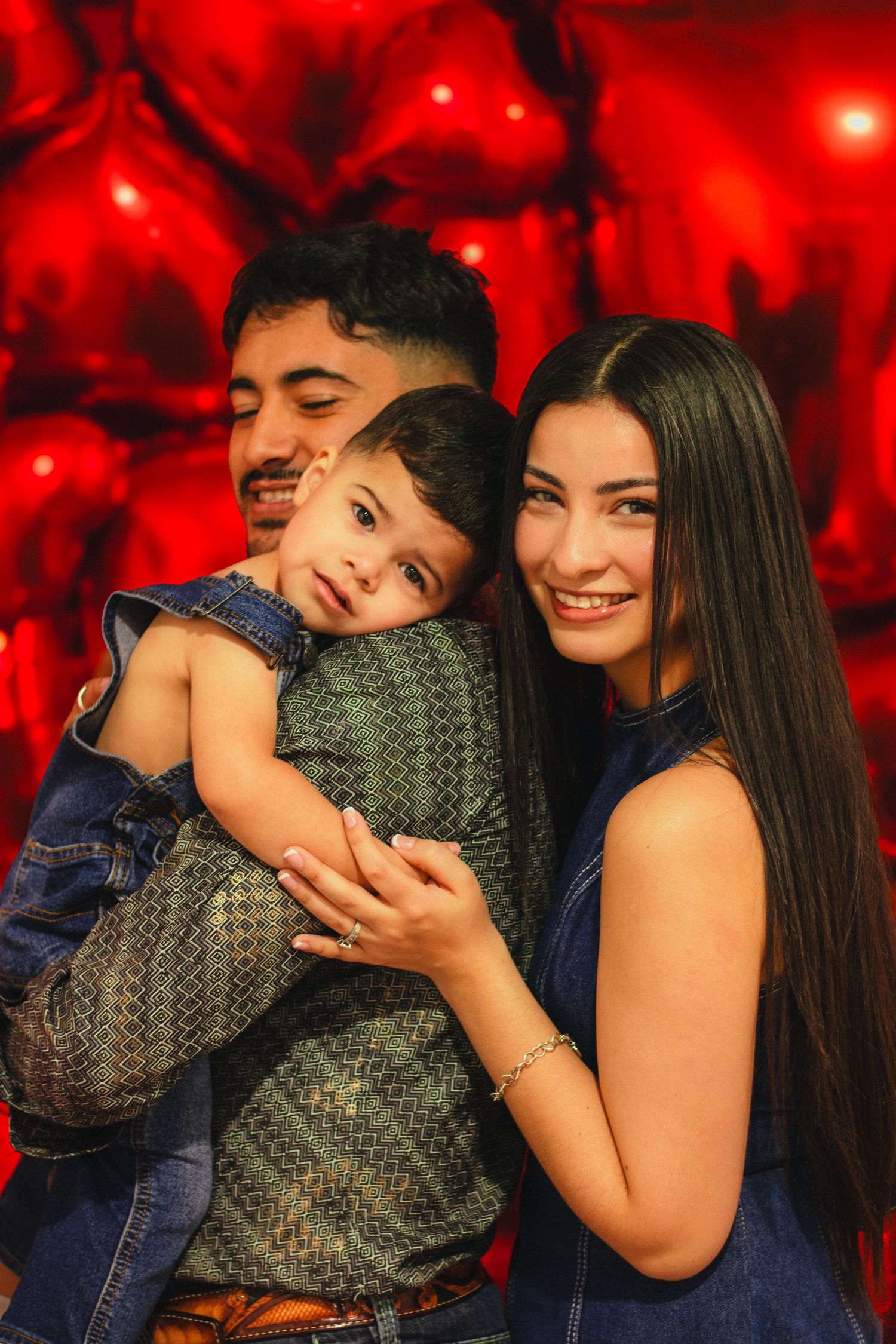 Happy young family with mother, father, and toddler posing together in front of red heart balloons.