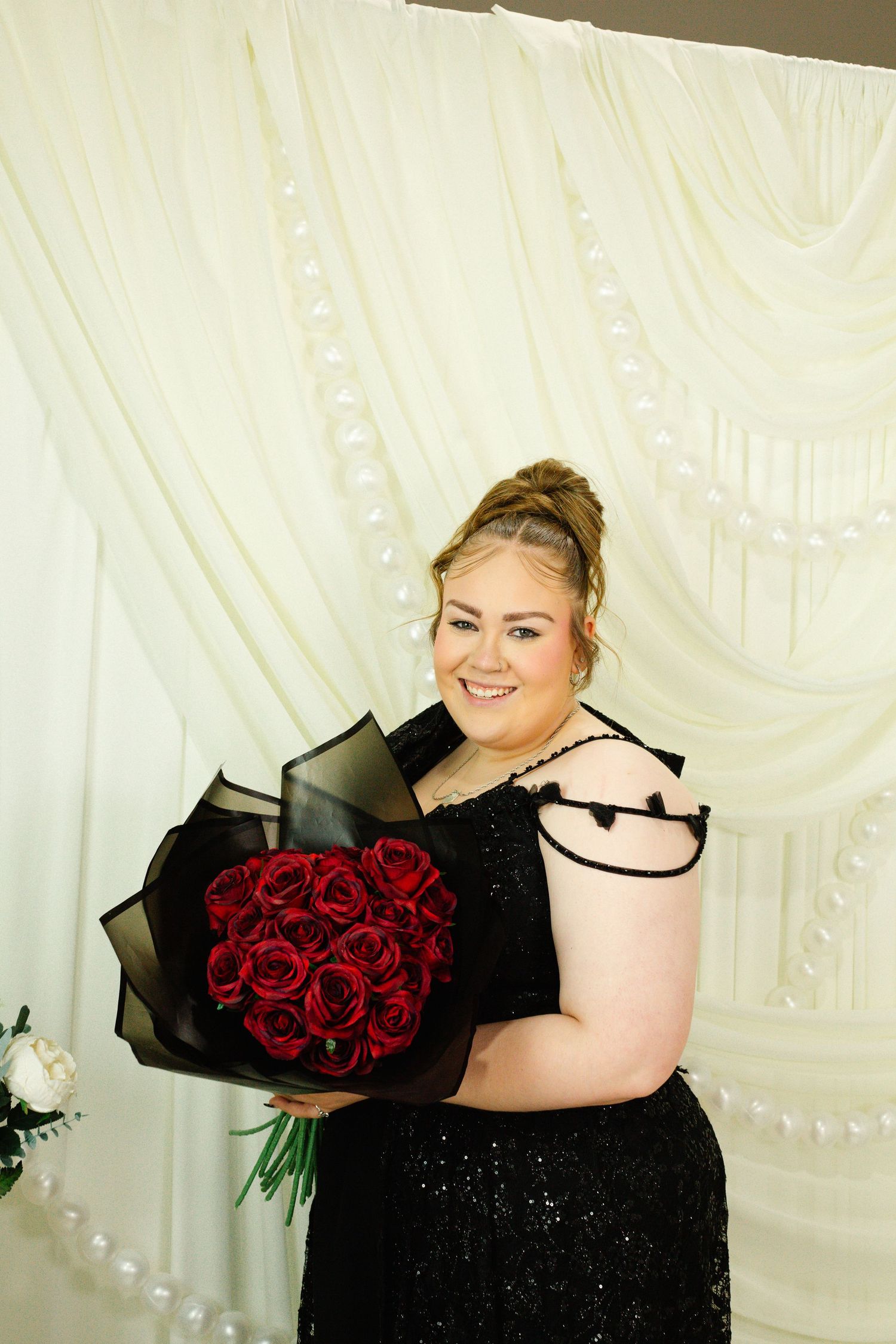 Woman in black sequin dress holding red roses bouquet, smiling in front of white draped backdrop.