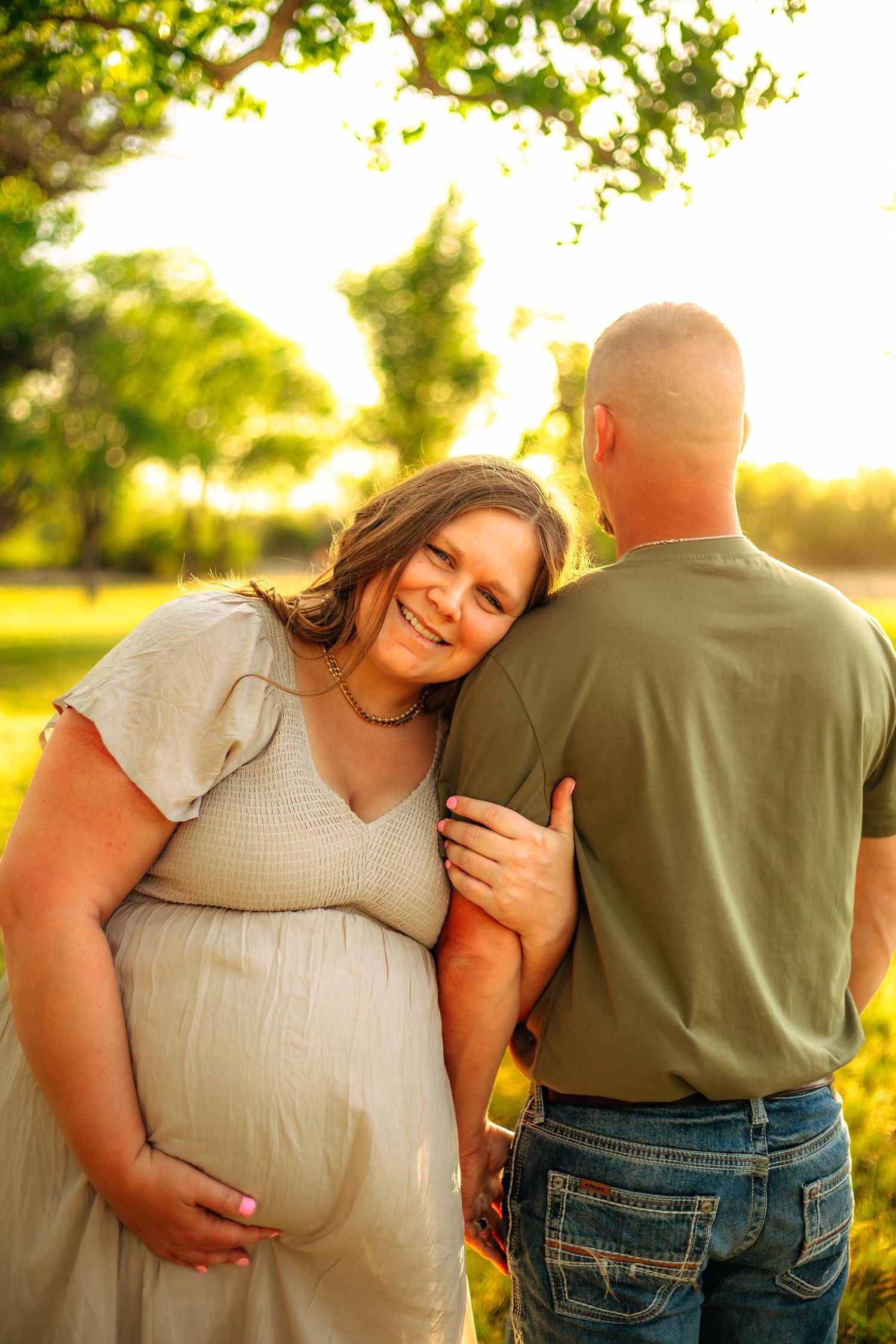 Pregnant woman in white dress smiles leaning on partner's shoulder during golden hour maternity photo outdoors.