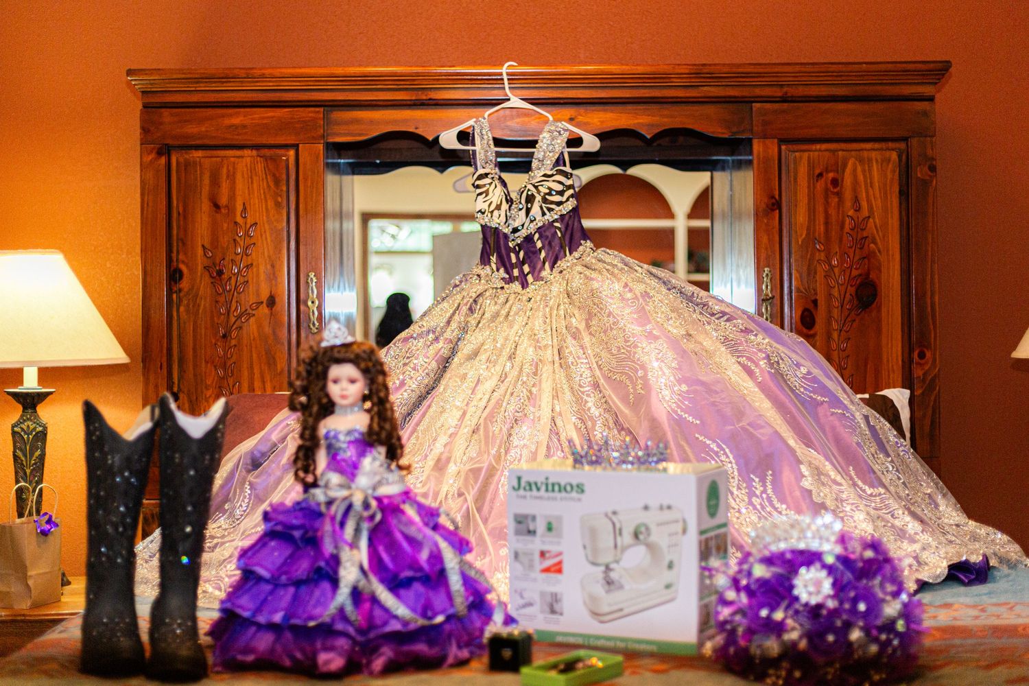 Quinceañera setup with gold ballgown, purple doll, cowboy boots, bouquet, and sewing machine on a wooden dresser.