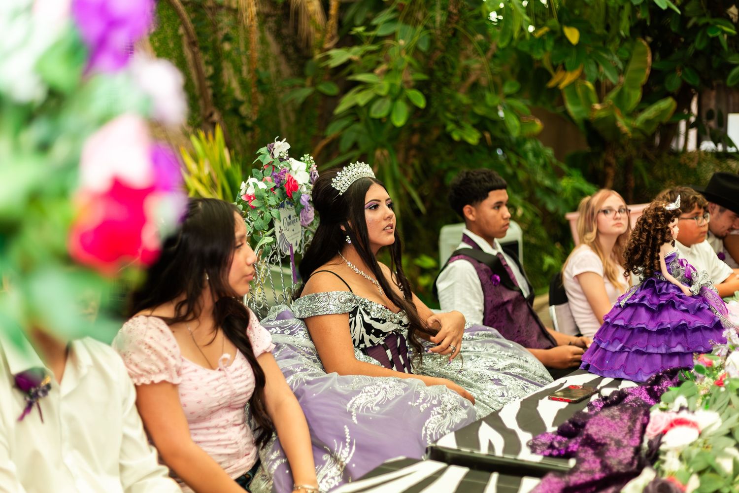 Young woman in elegant quinceañera gown with floral crown seated outdoors among guests at a colorful celebration.