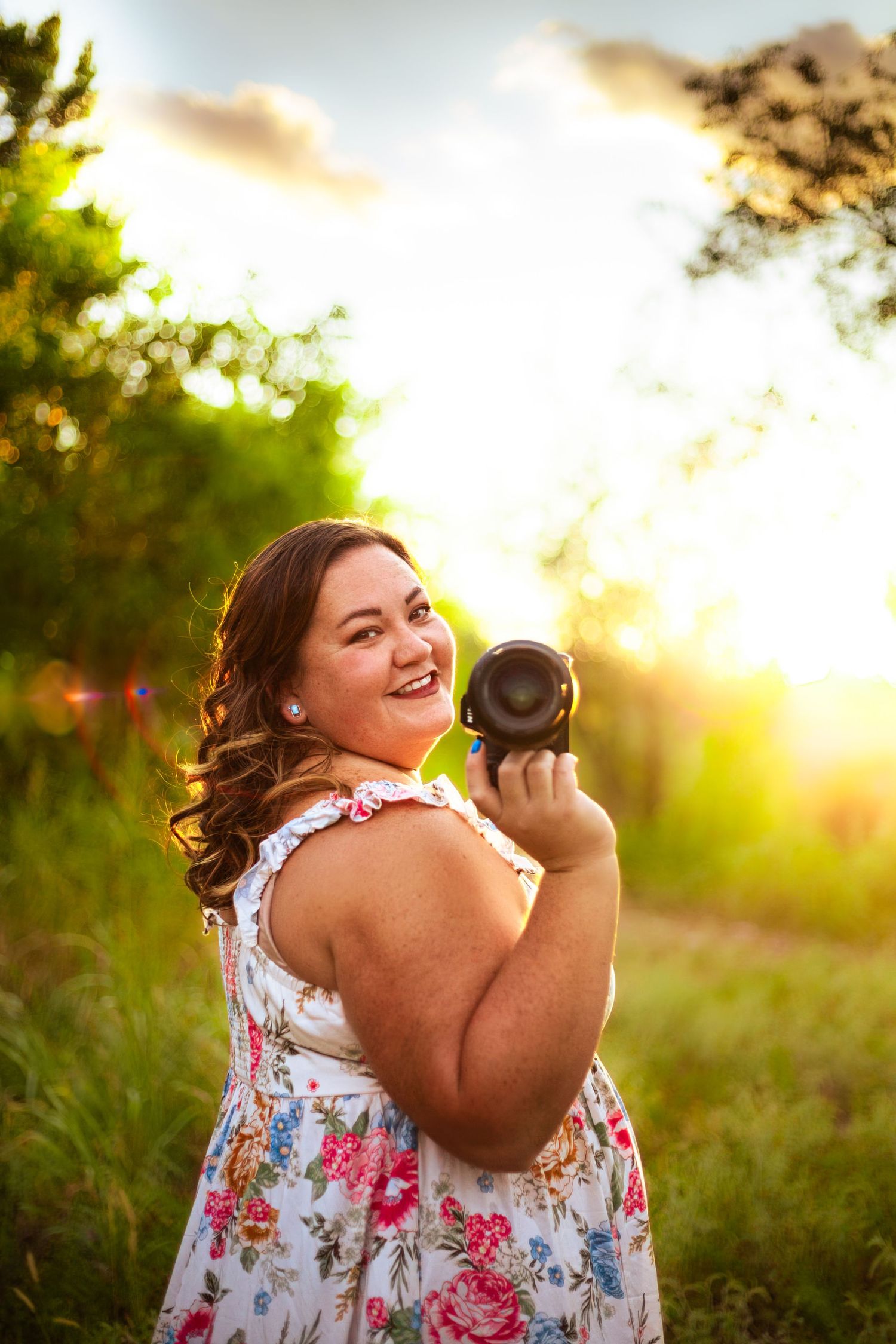Smiling woman in floral dress holding a camera outdoors at golden hour with lush green trees in background.