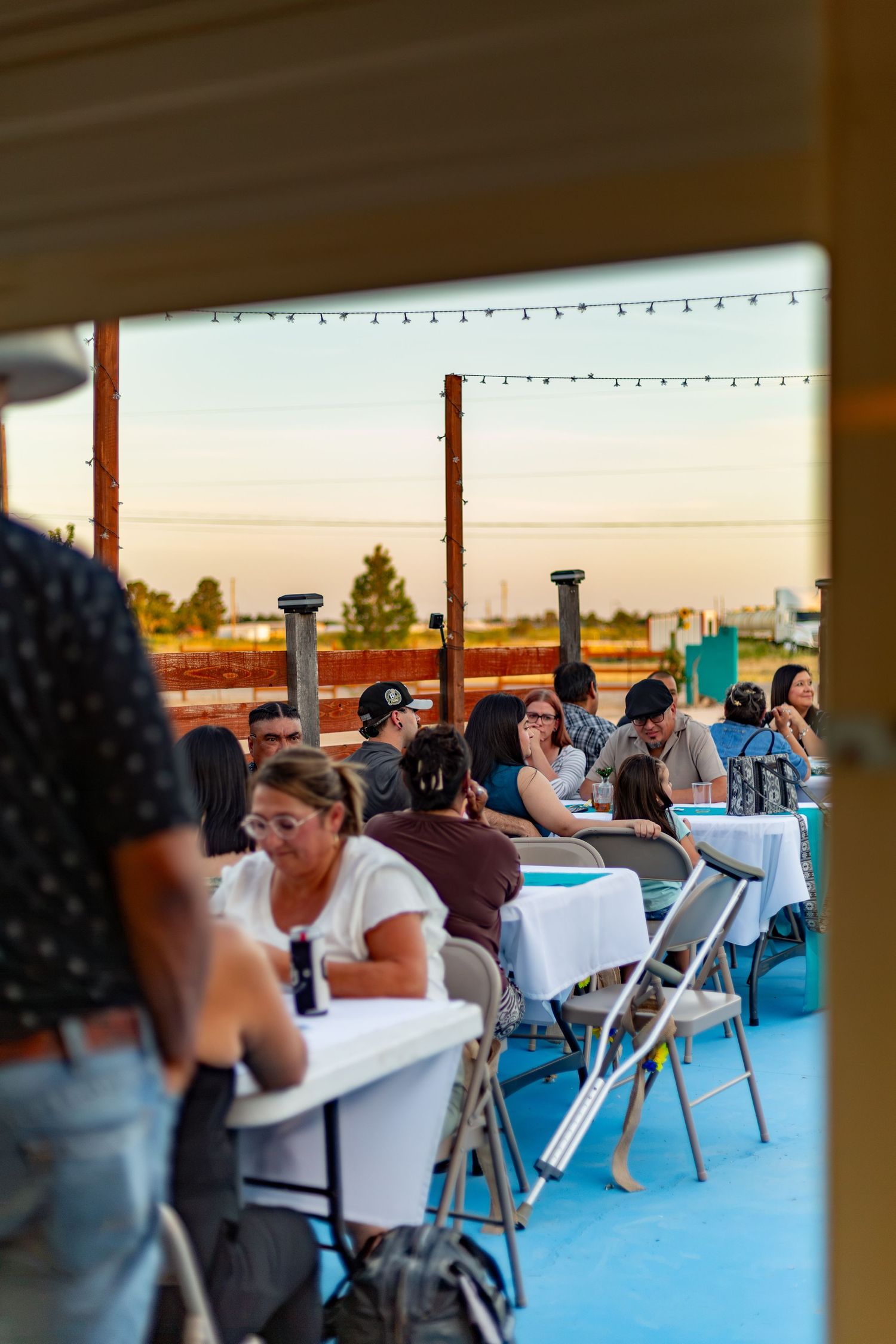 Wedding reception guests dining at white-clothed tables on outdoor patio at sunset.