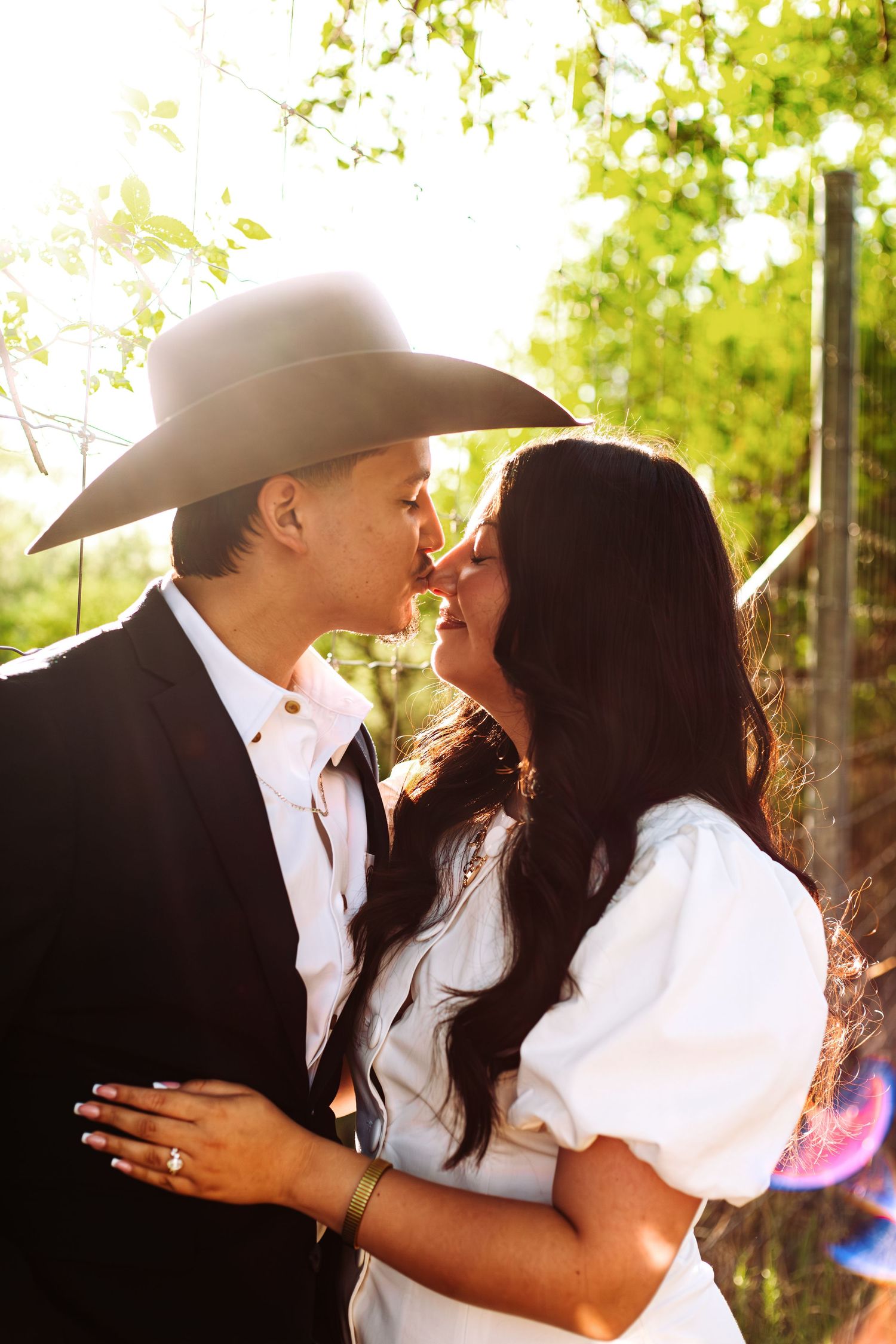 Couple sharing a romantic kiss outdoors, man in black suit and cowboy hat, woman in white dress, golden sunlight.