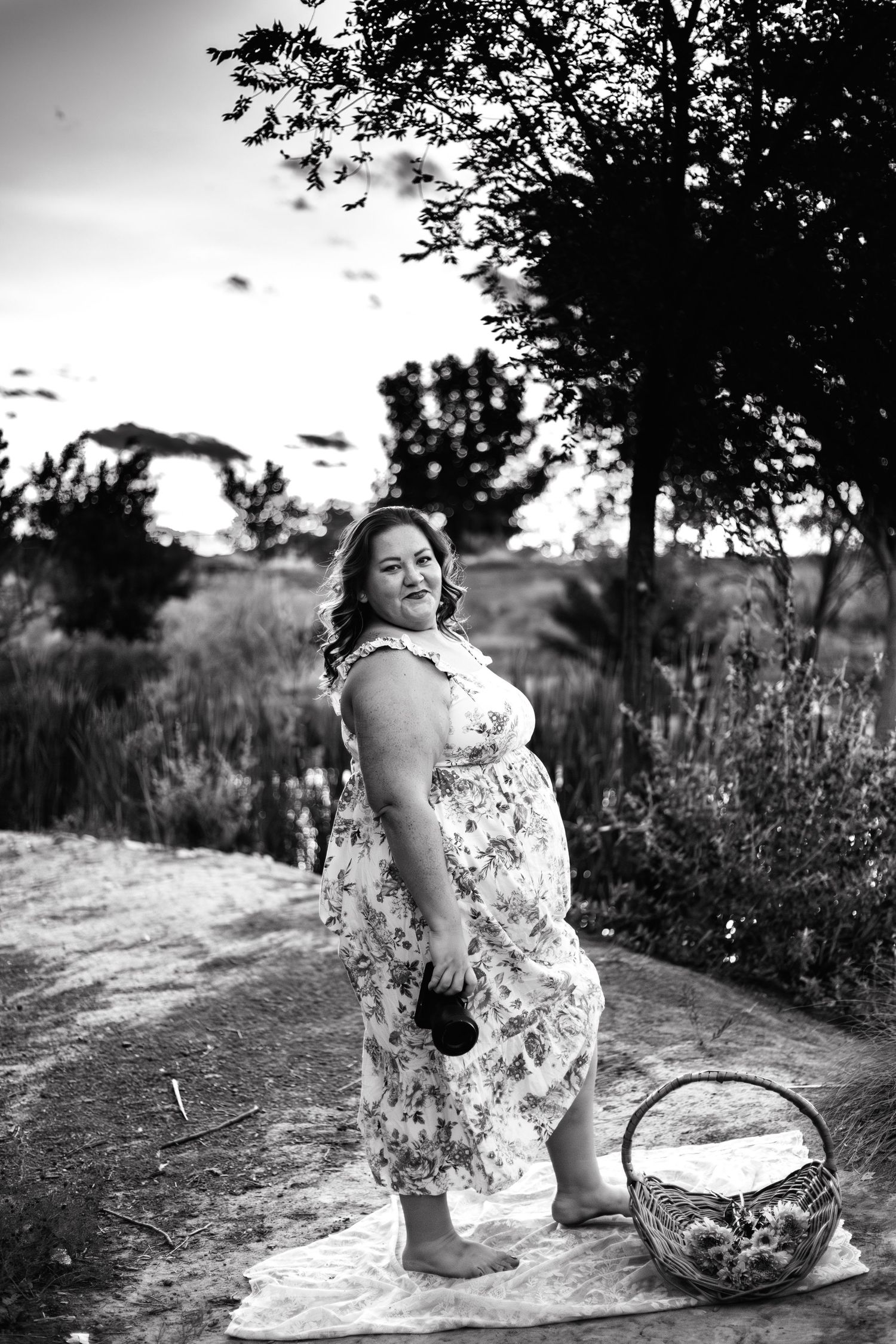 Black and white photo of a pregnant woman in a floral dress standing barefoot outdoors near a wicker basket.