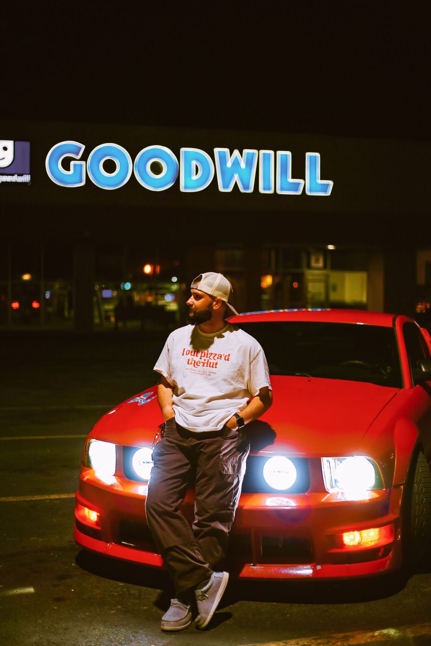Man in white tee and cap leans on red Mustang at night under a glowing Goodwill sign.