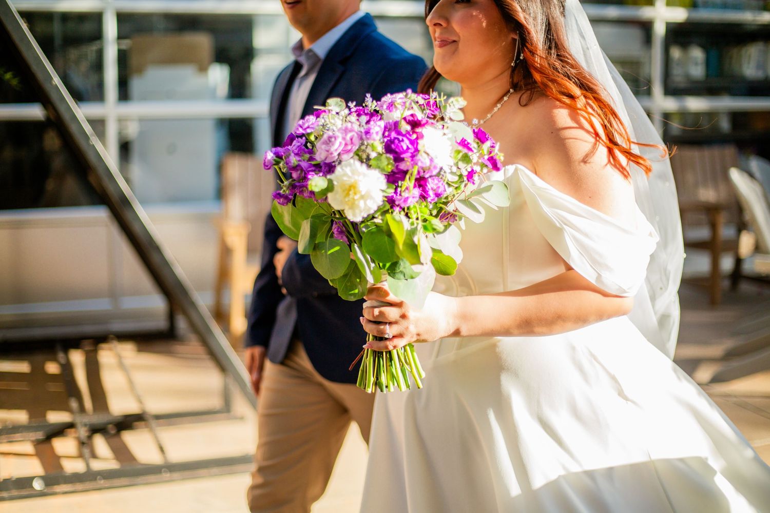 Bride in white off-shoulder dress holding purple and white wedding bouquet with groom in blue suit.